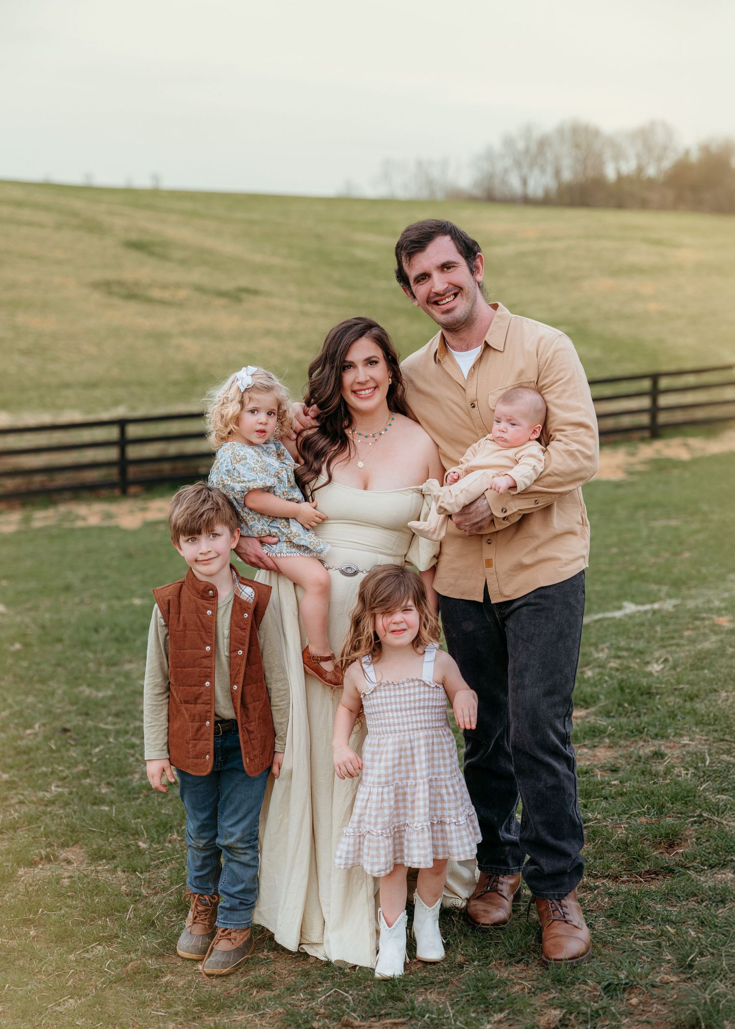 A family group poses together in a grassy field on a sunny day wearing coordinated brown and white outfits.