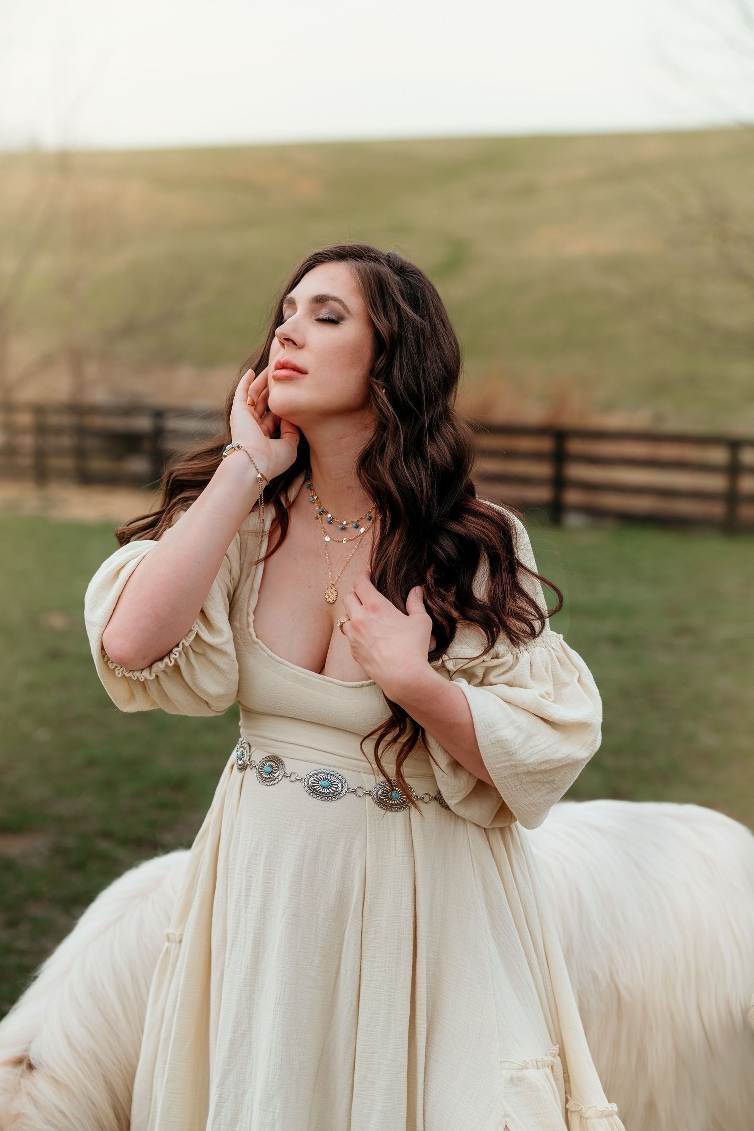 A dreamy bridal portrait in a cream-colored dress with billowing sleeves against a pastoral countryside backdrop.