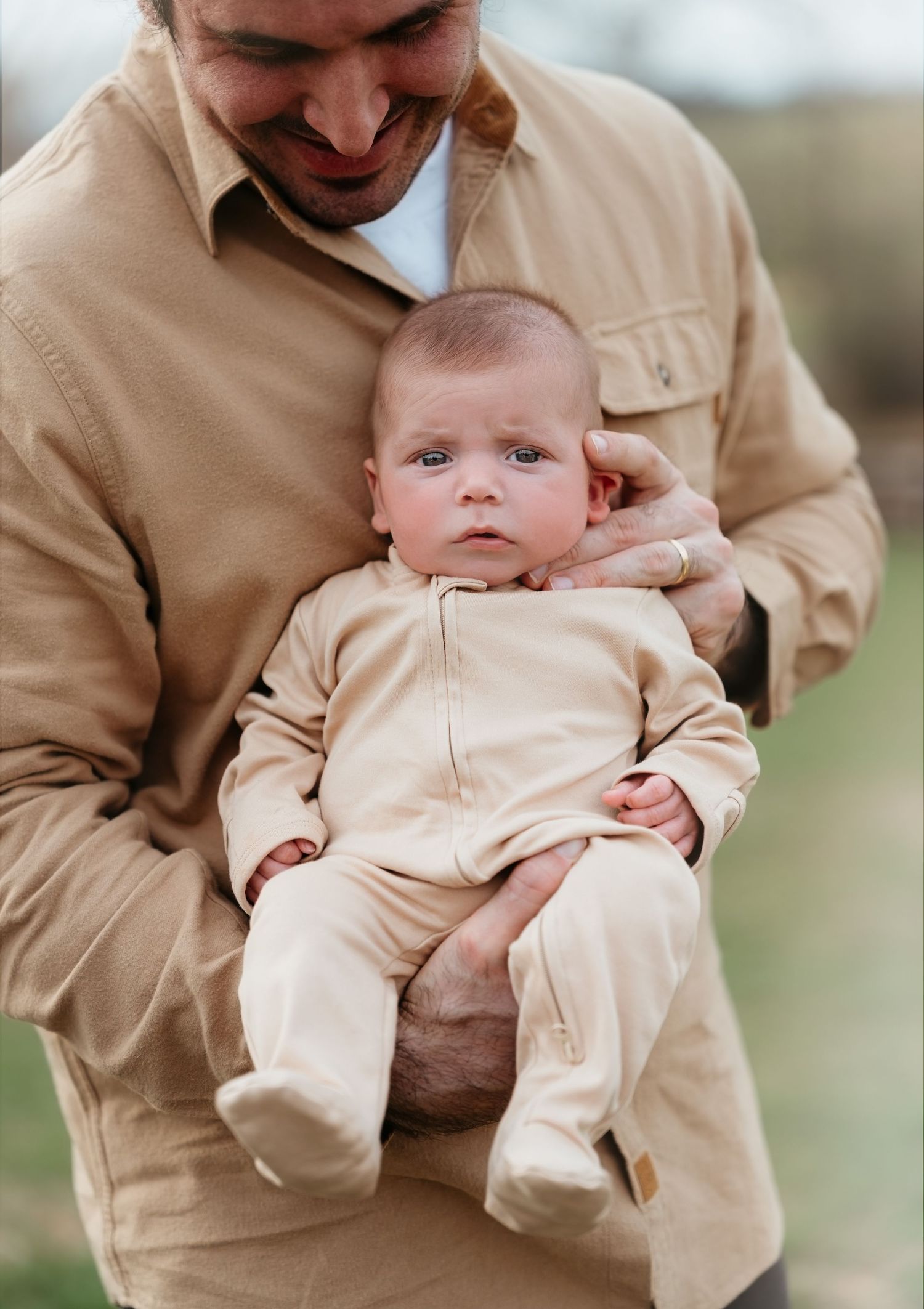 A baby in a beige onesie is held lovingly in someone's arms while outdoors.