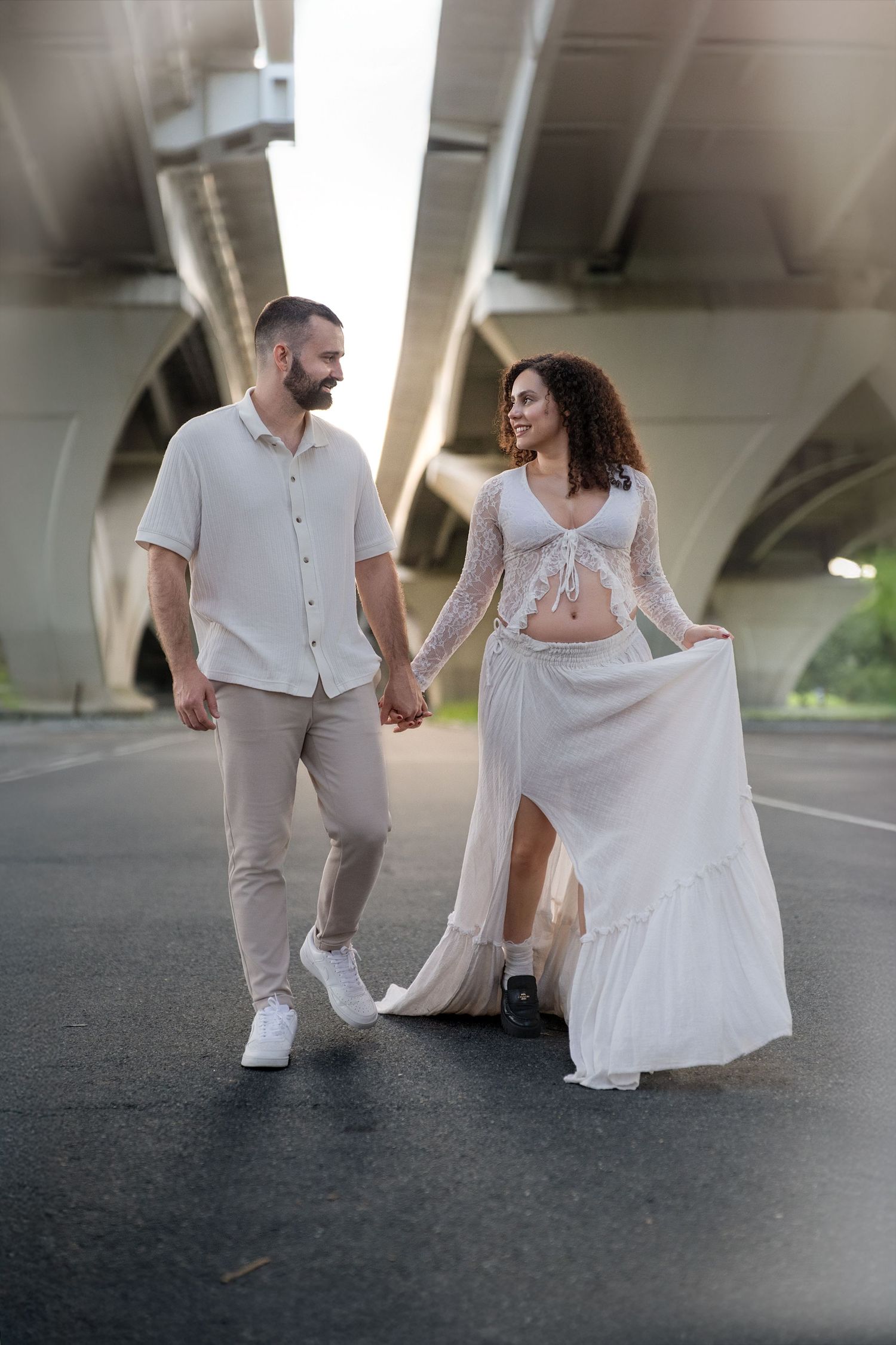 Artistic maternity photos under bridge with couple in white and beige outfits.
