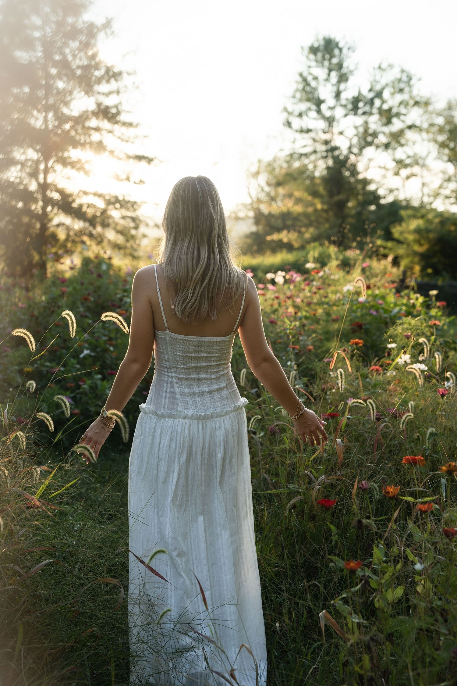 Woman in white dress walking through wildflower garden at golden hour sunset.