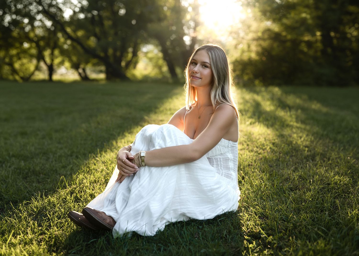 Young blonde woman in white dress sitting on lush green grass during golden hour sunset in park.