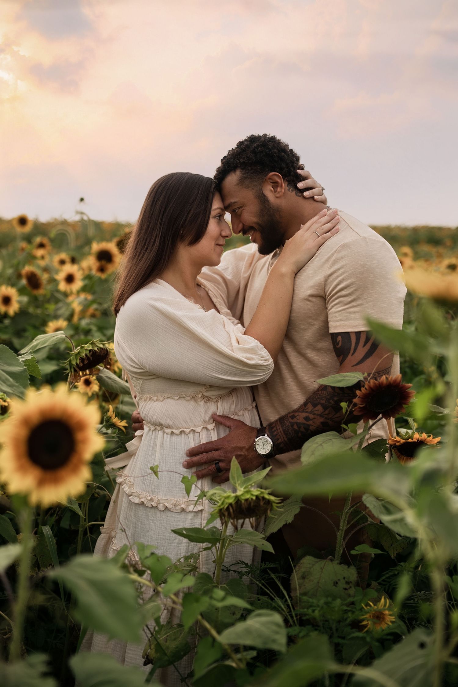 Romantic maternity photos in sunflower field at sunset with couple in light-colored clothing.