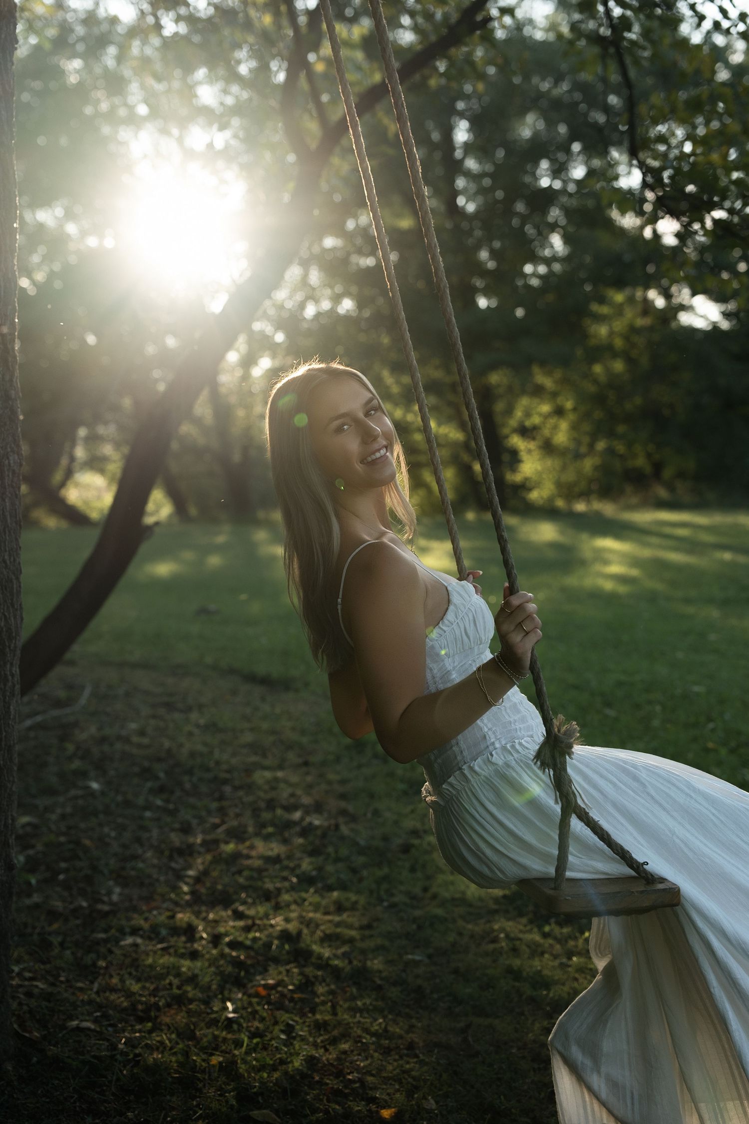 Young woman in white dress smiling on a rope swing in a sunlit green park.
