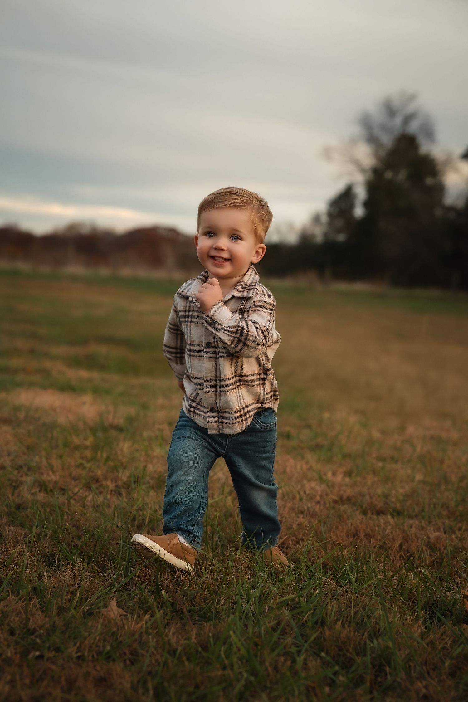 A toddler in a plaid shirt and jeans plays in a grassy field during golden hour.