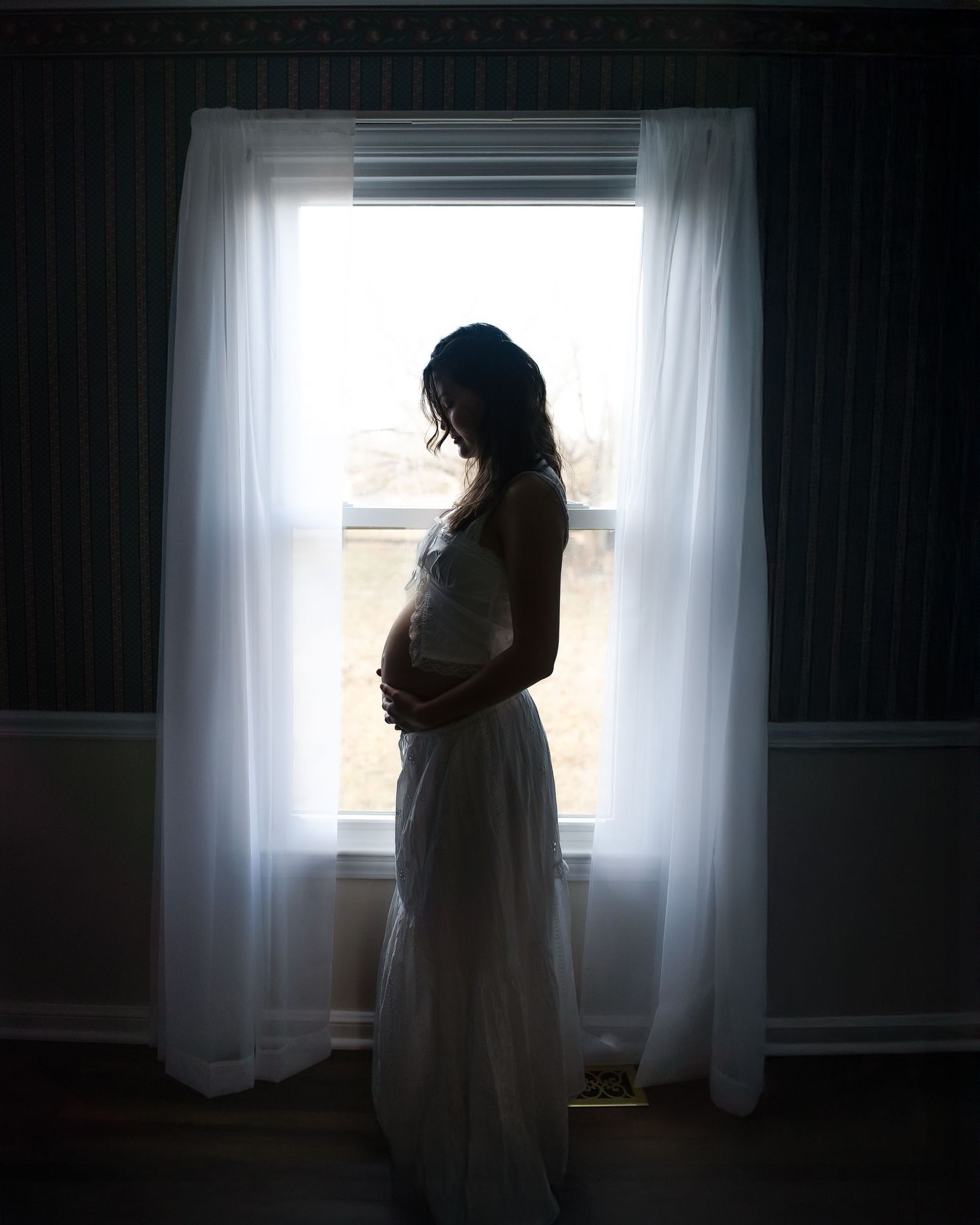 Silhouette of an expectant mother in a white dress standing by a sunlit window with flowing curtains.