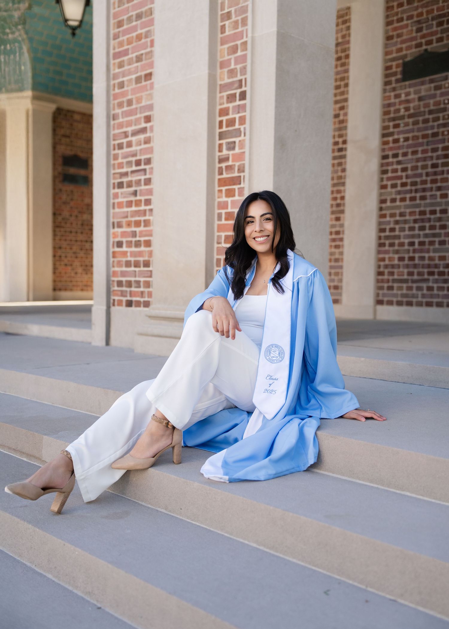 A smiling graduate sits on steps wearing a light blue graduation gown and white sash outdoors.