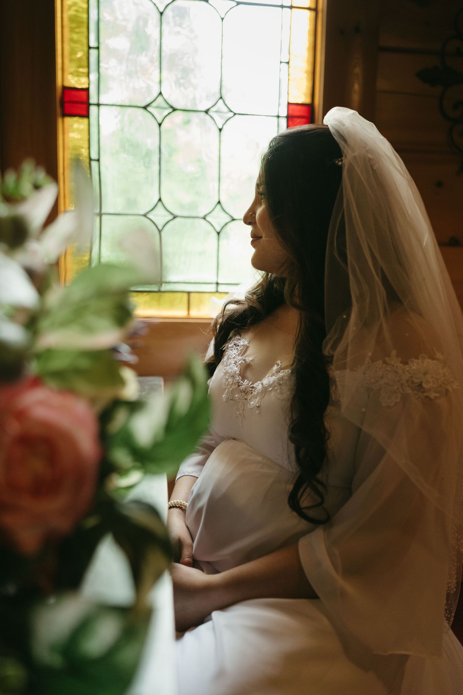 A bride in white lace gown and veil poses by stained glass window with roses in foreground.