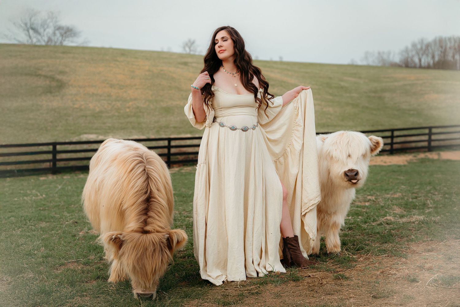 A woman in a flowing cream dress poses with a majestic golden retriever dog in a rustic farm setting at sunset.