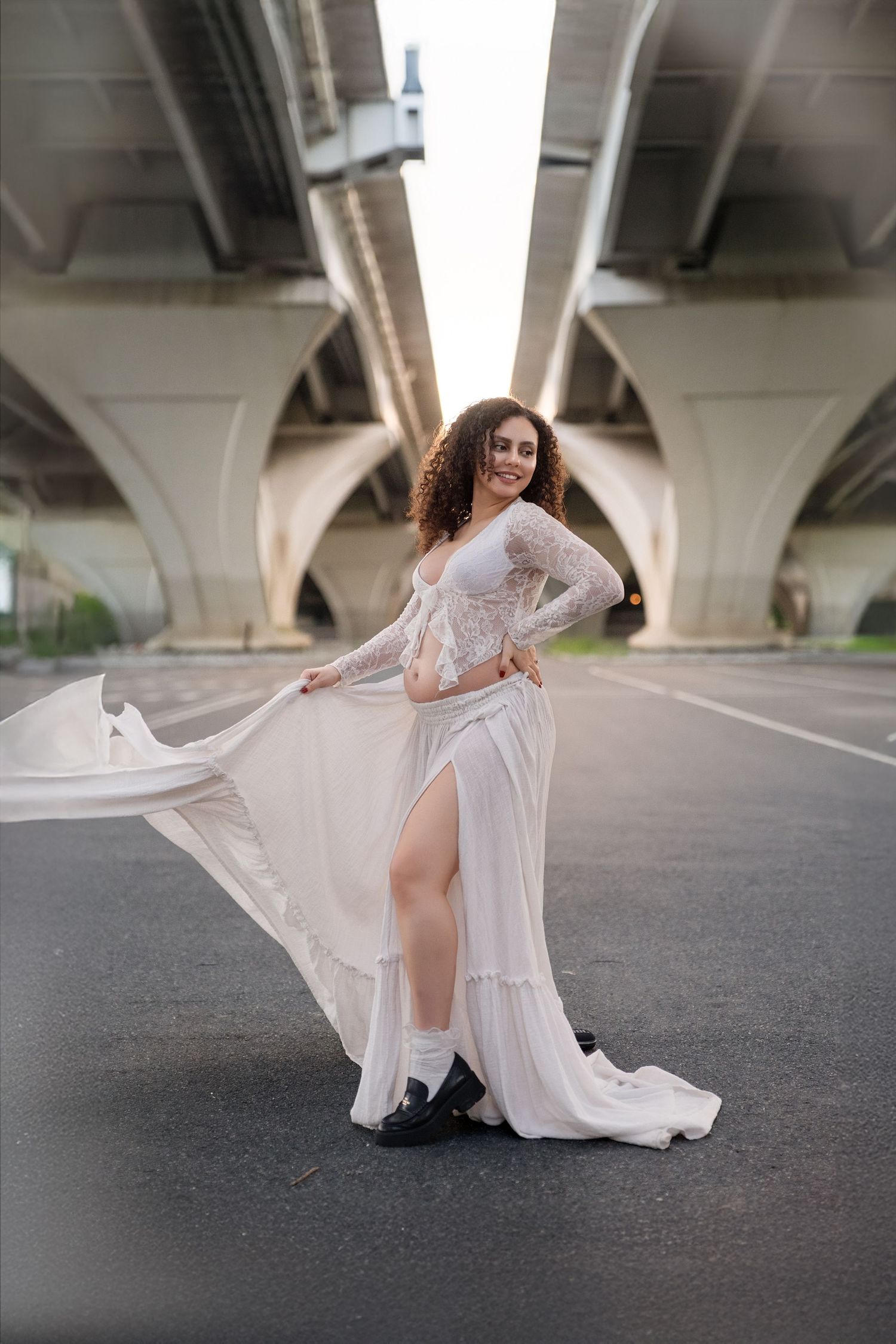 Elegant maternity shoot showcasing flowing white lace dress under architectural bridge.