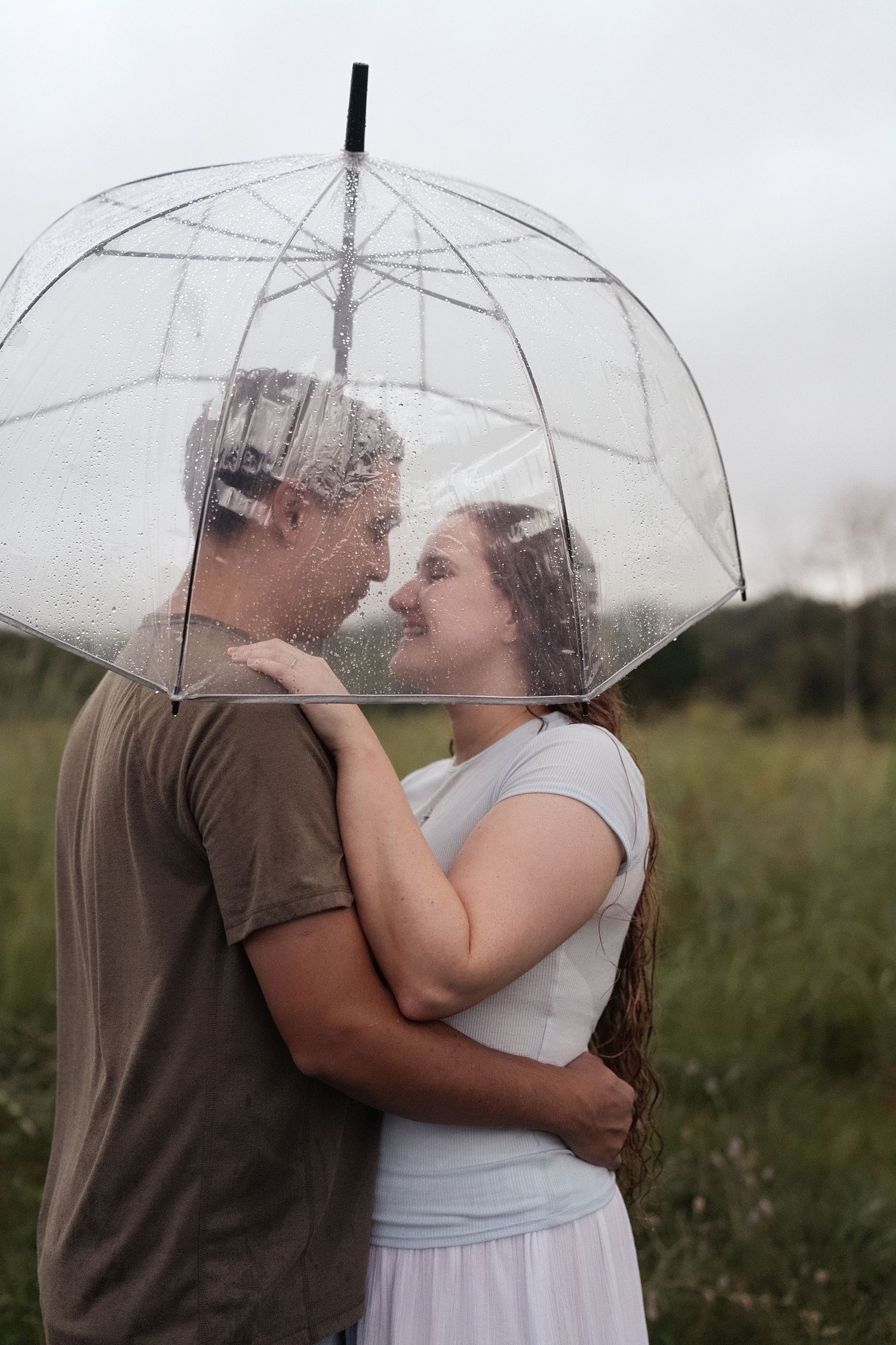 Couple embracing under a clear bubble umbrella in a rainy field, sharing a romantic moment.