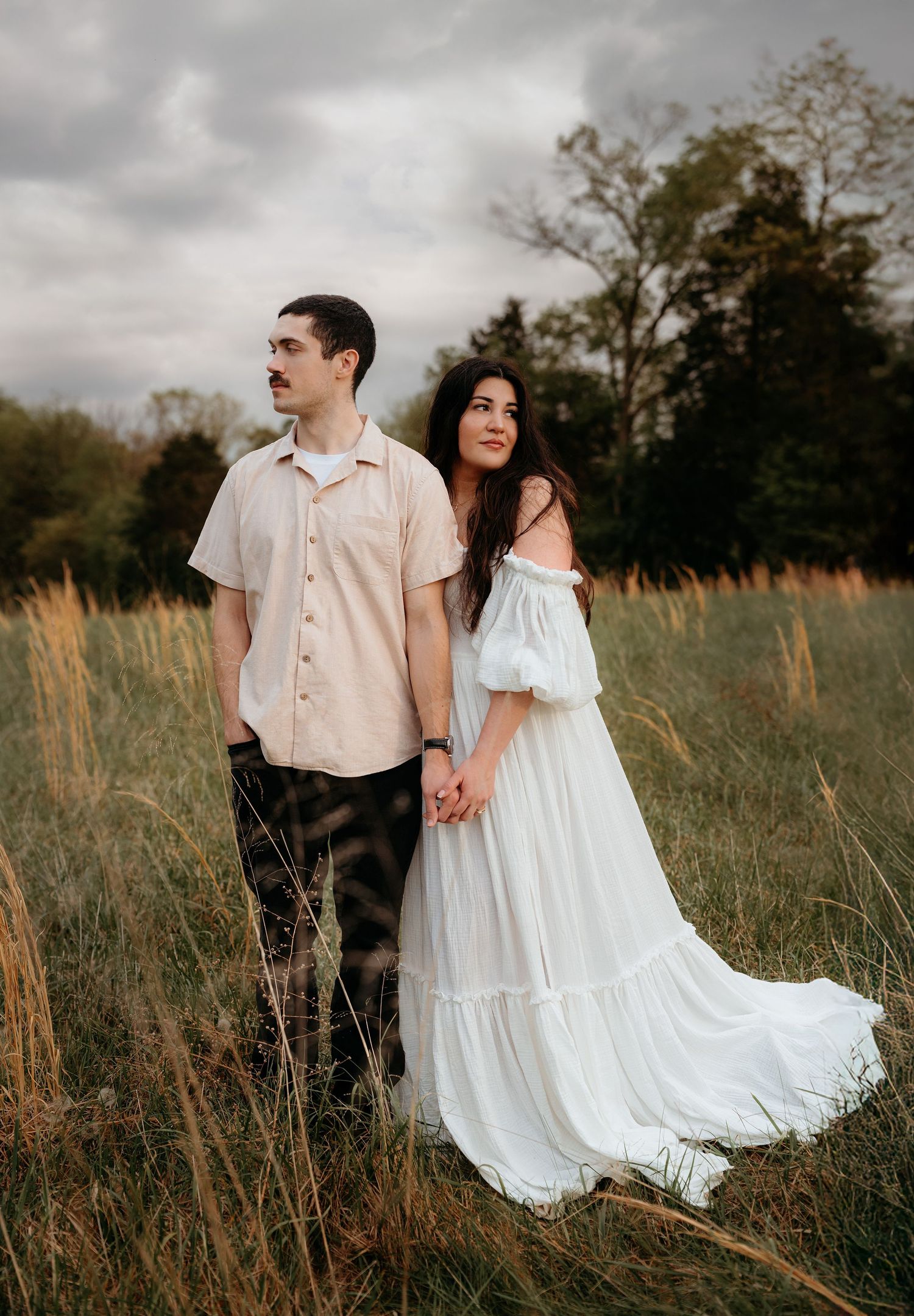 A couple holds hands while walking through a field of tall grass under moody skies, with her white dress flowing.