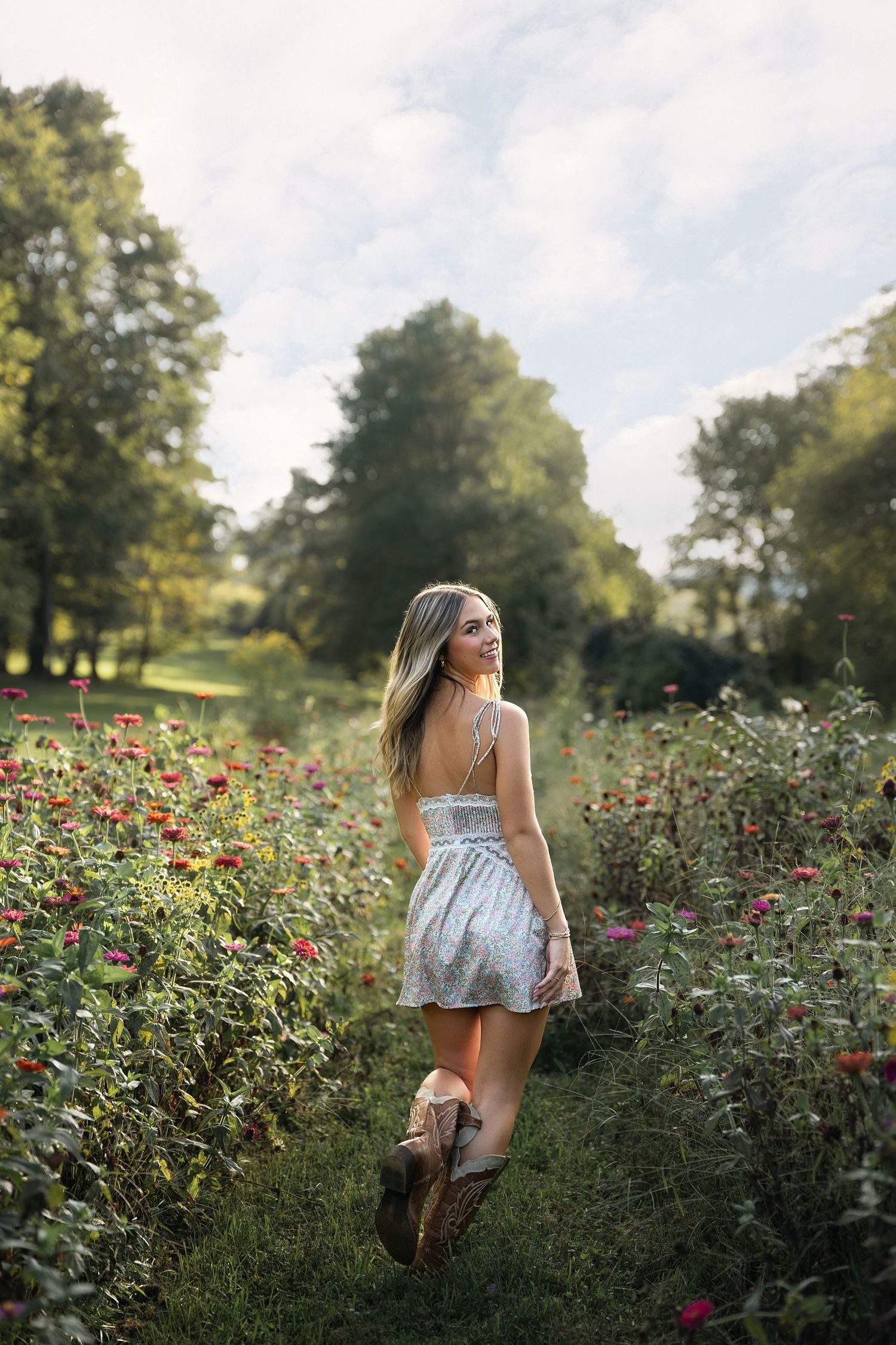 Young woman in white sundress and boots walks through a vibrant flower garden path surrounded by lush green trees.