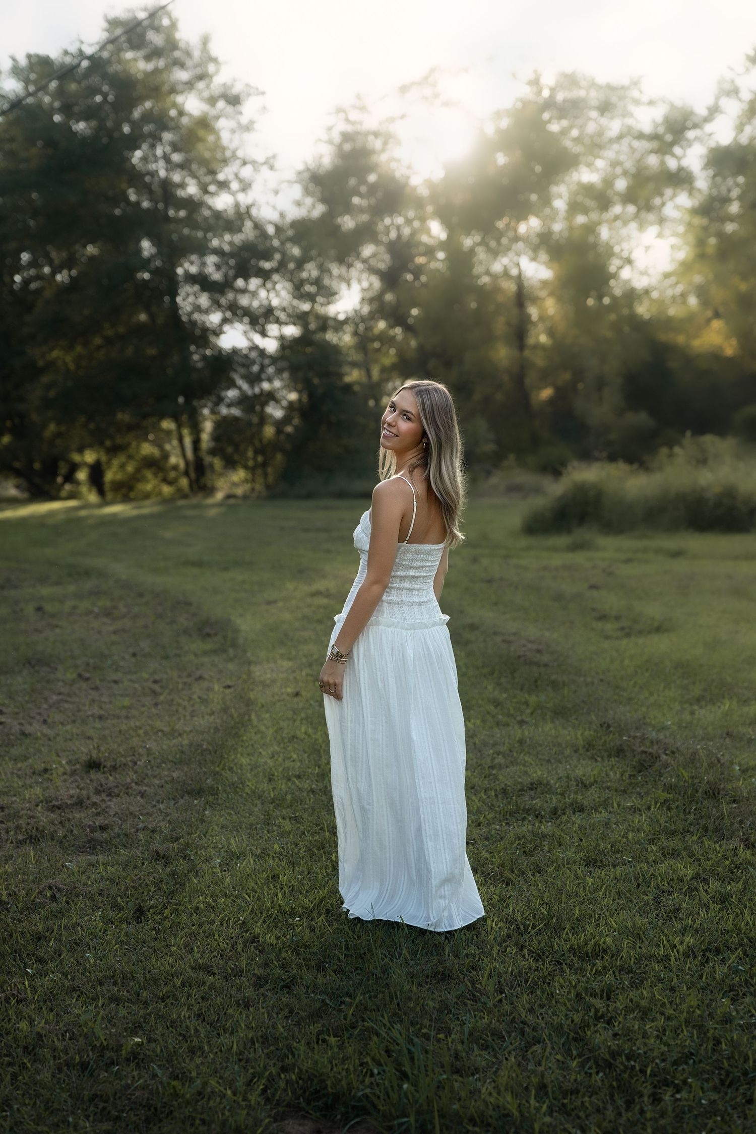 Young woman in white maxi dress standing on green grass field at golden hour sunset with trees in background.