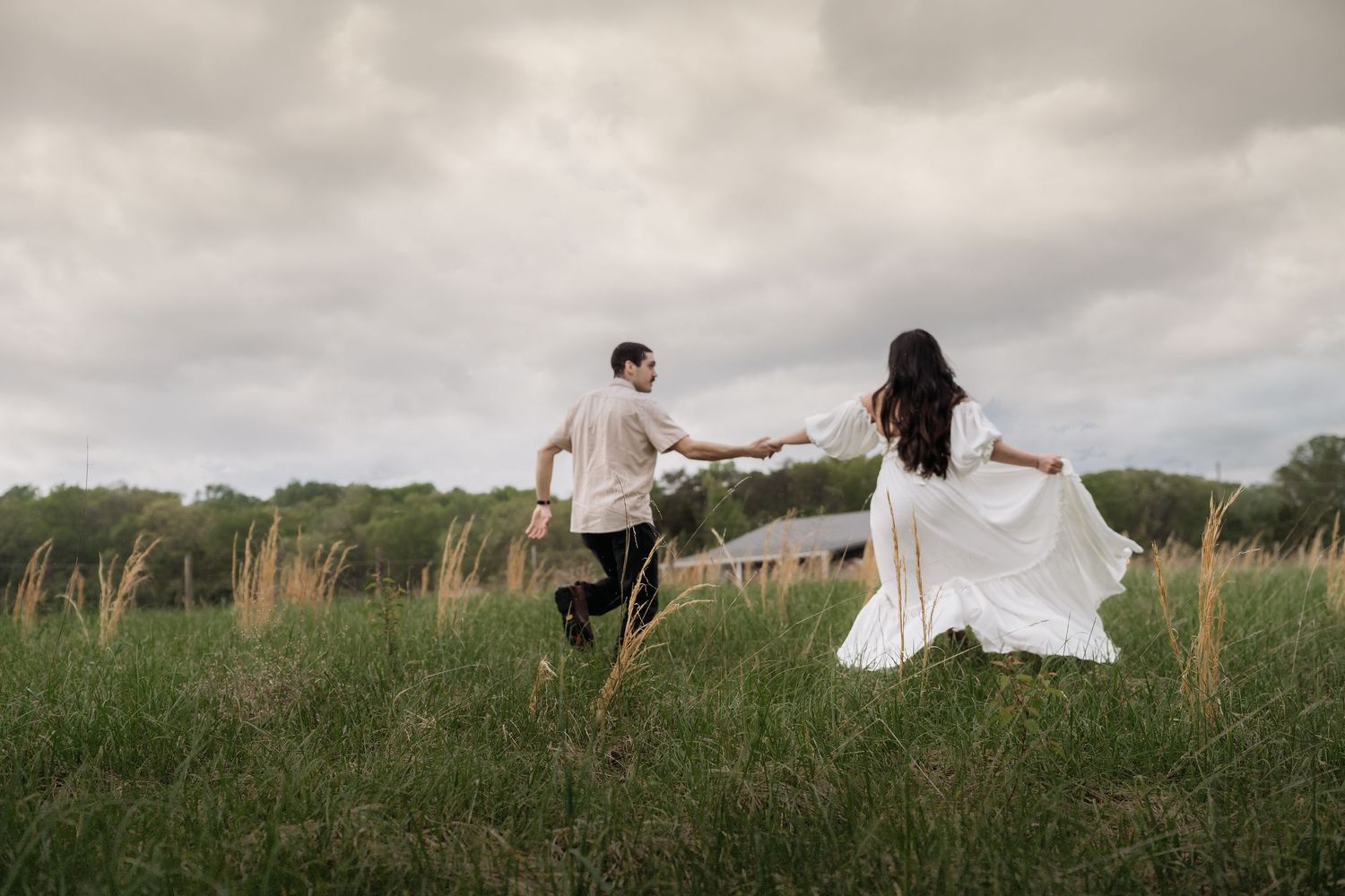 Couple playfully running through a grassy field at golden hour with trees in the background.