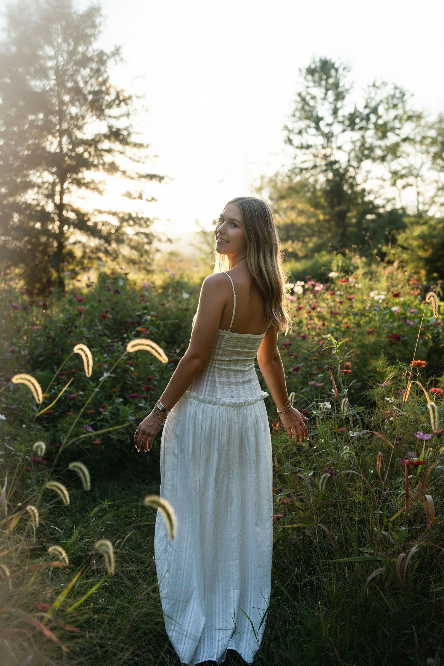 Woman in white maxi dress standing in a sunlit wildflower garden surrounded by lush greenery at golden hour.