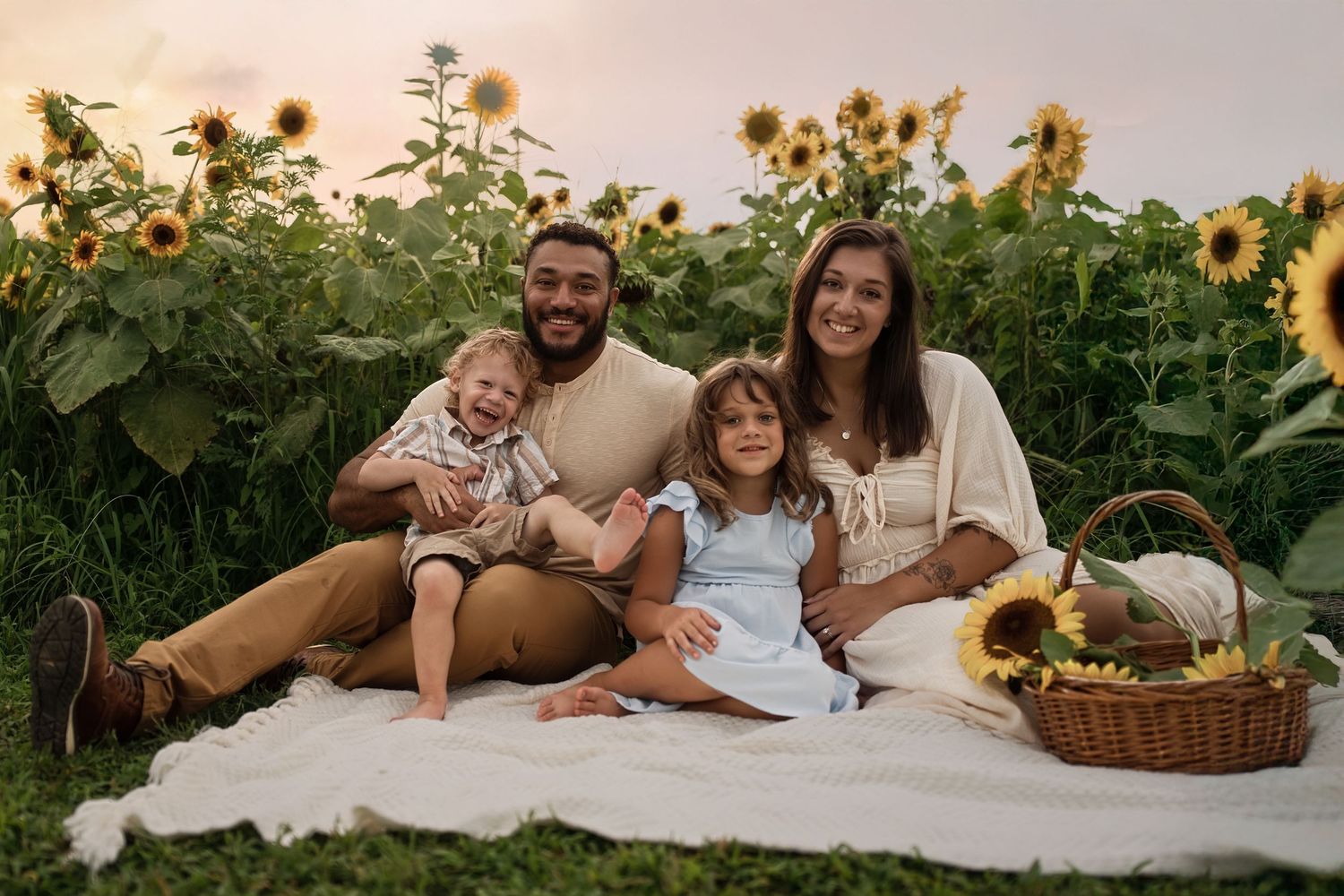 A family sits on a white blanket in a sunflower field at sunset with a picnic basket beside them.