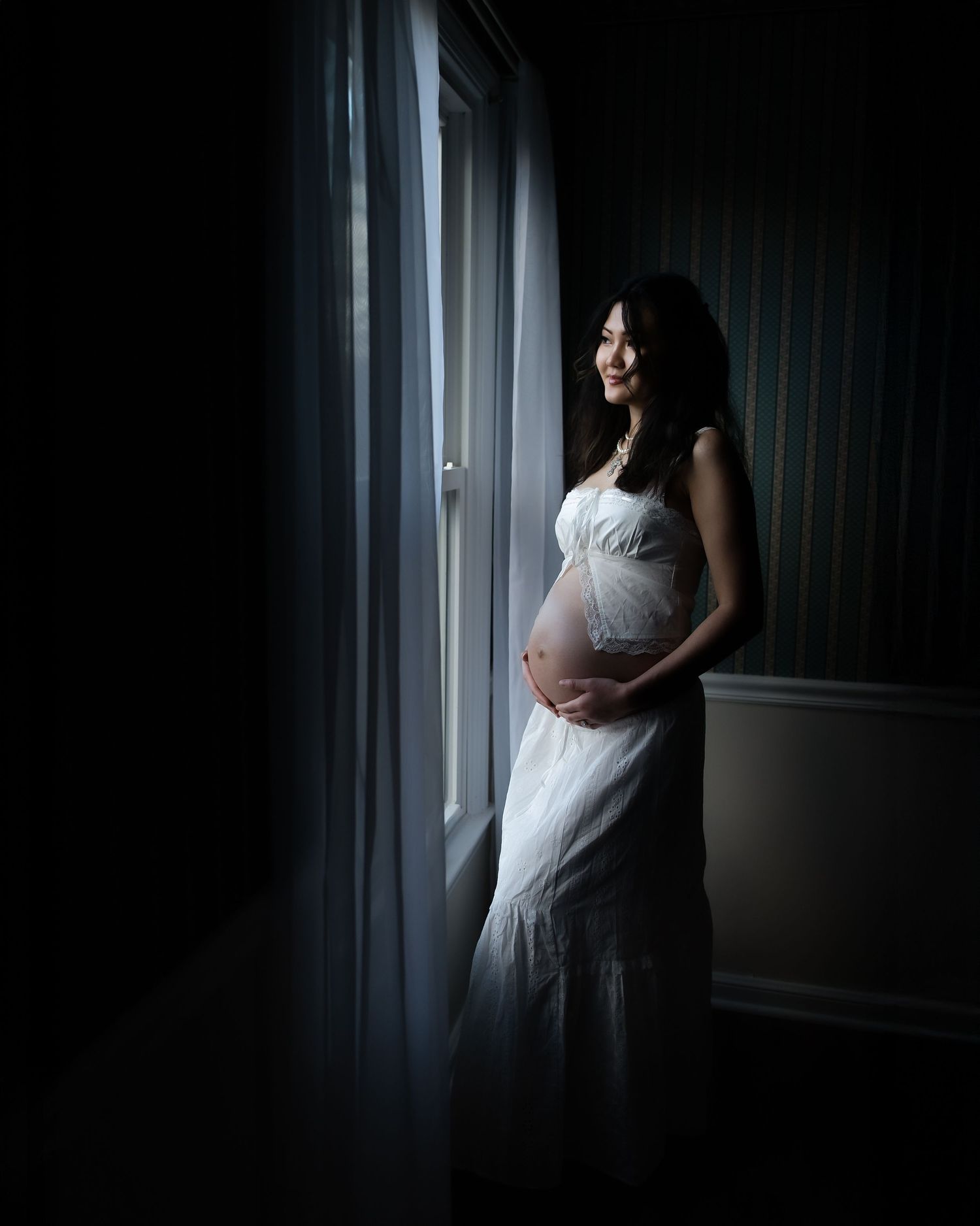 Moody maternity photos in dark room with dramatic window lighting creating silhouettes.