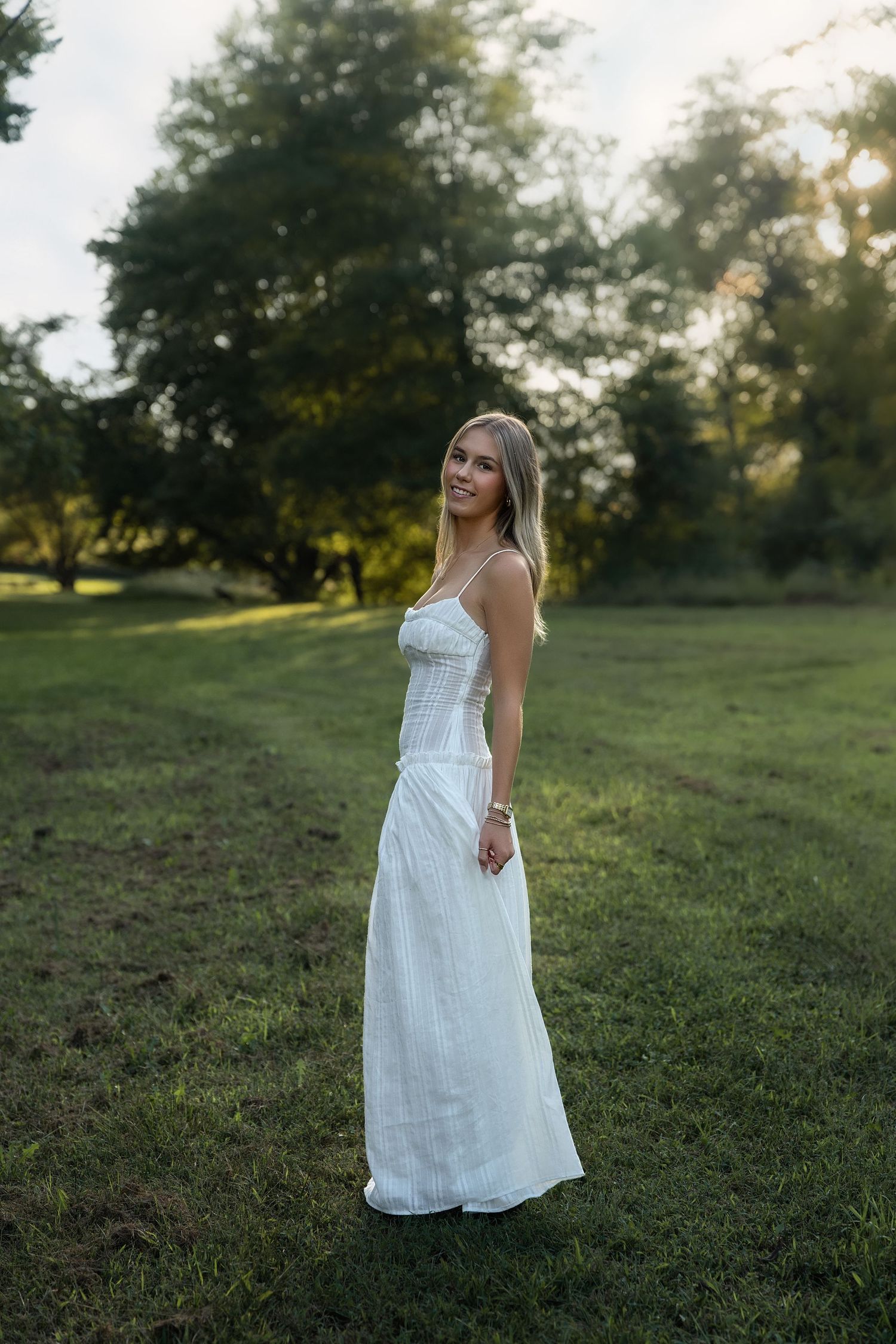 Young woman in white maxi dress standing on green grass field with trees in golden hour sunlight.