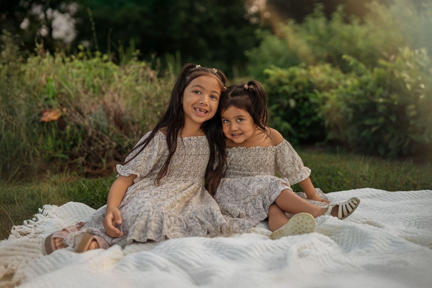 Two people in matching floral dresses share a tender moment on a white blanket outdoors.