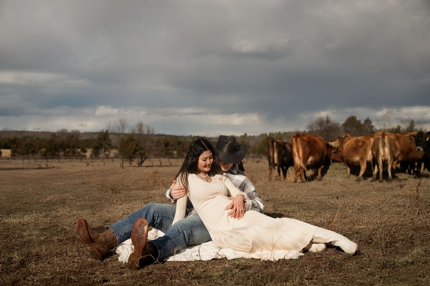 Couple relaxes on white blanket in field with horses grazing in background under cloudy sky.