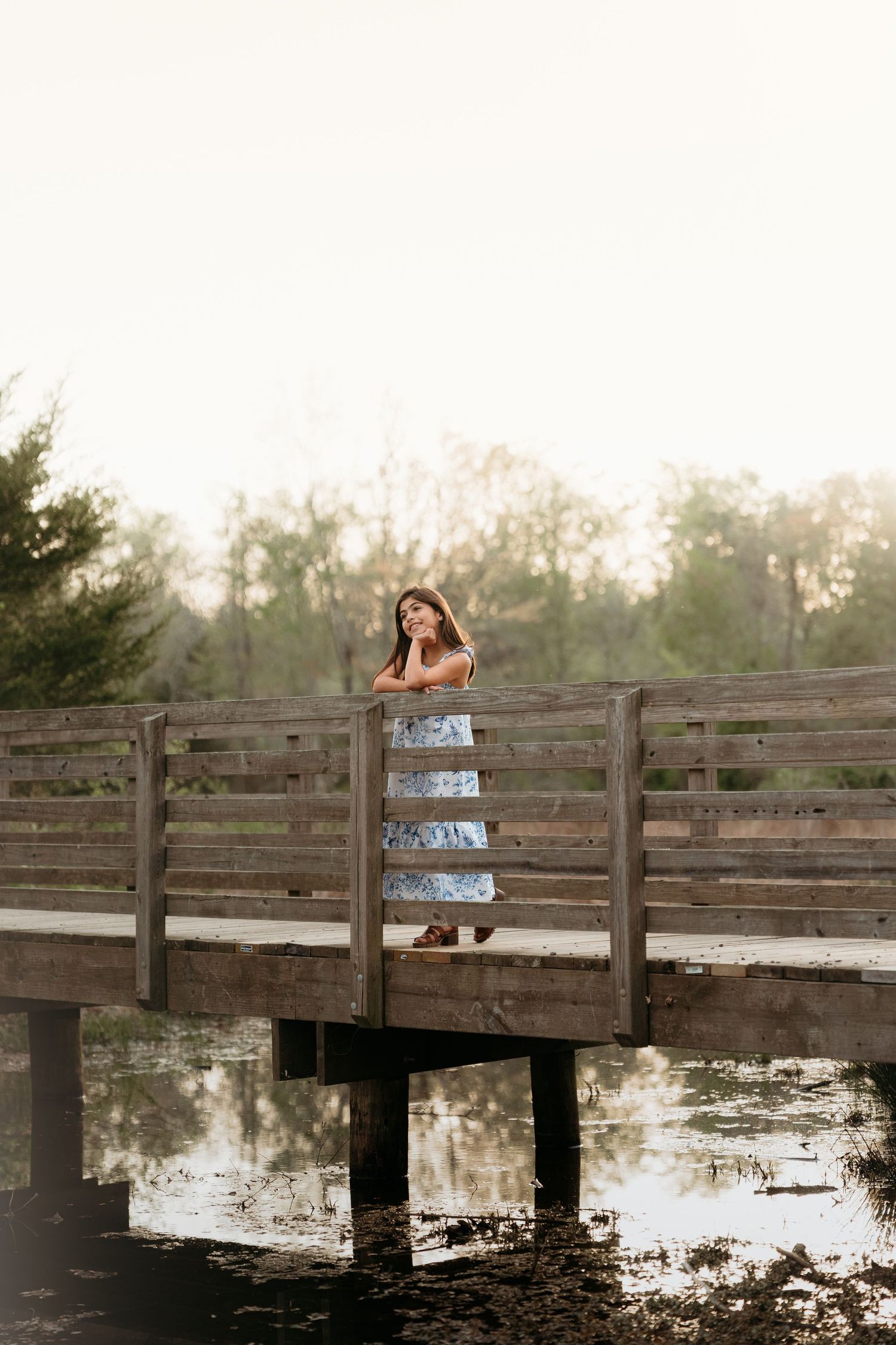 A wooden bridge stretches over a tranquil pond at sunset while a beautiful young lady in a blue dress poses on the railing in haymarket VA