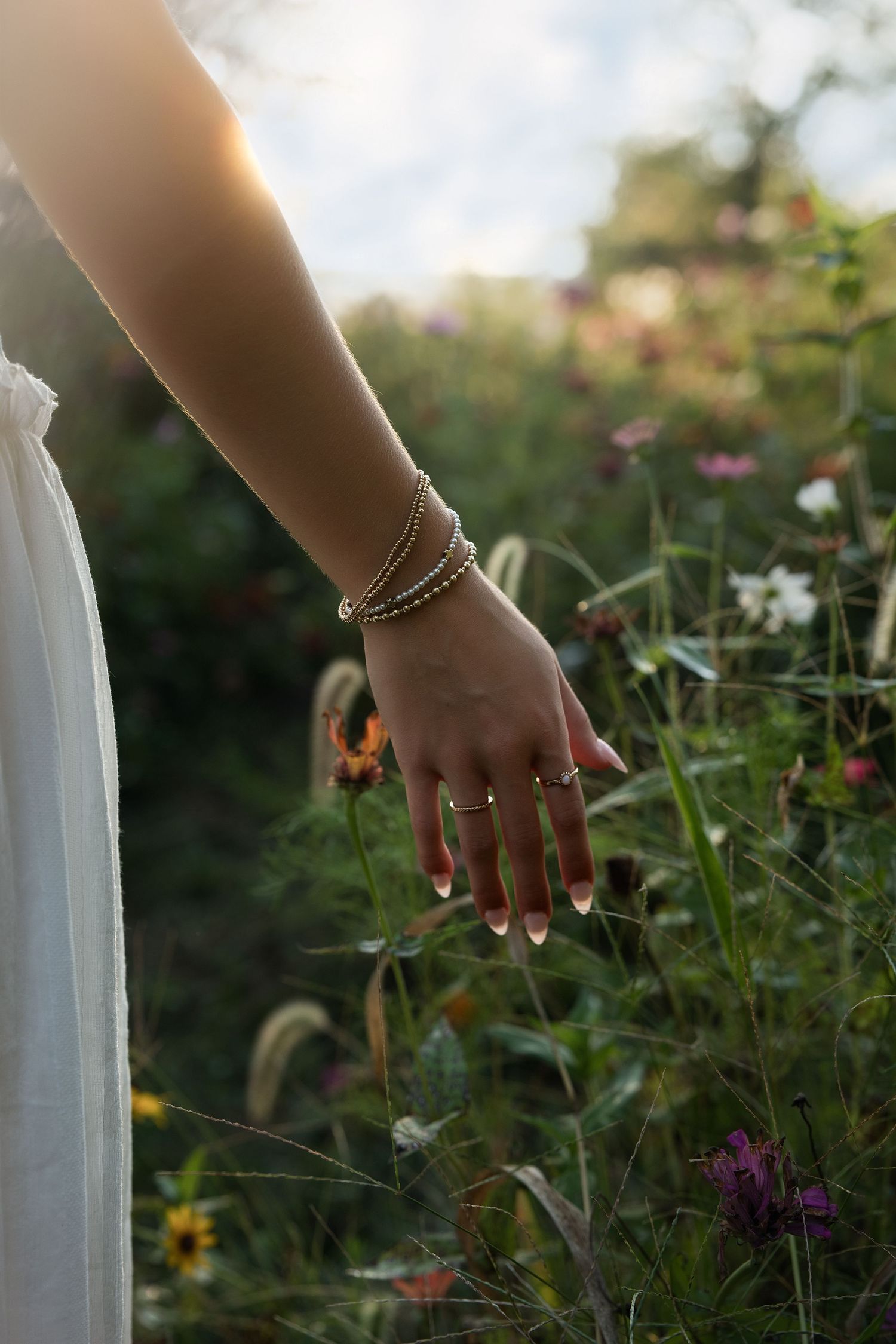 Woman's hand adorned with gold bracelets and rings grazing wildflowers in a sunlit garden.