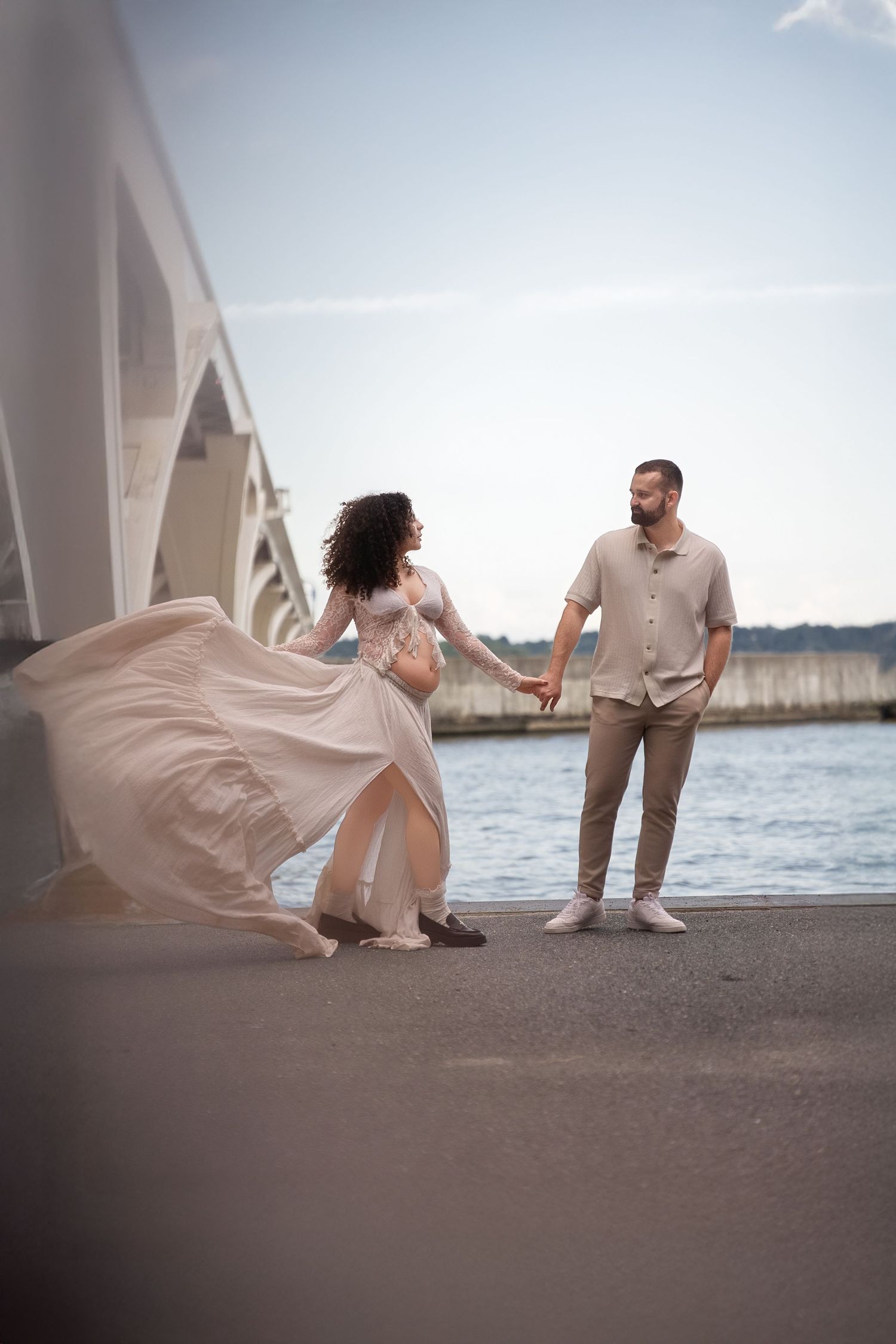 Romantic maternity photoshoot under a bridge with flowing white dress and water backdrop.
