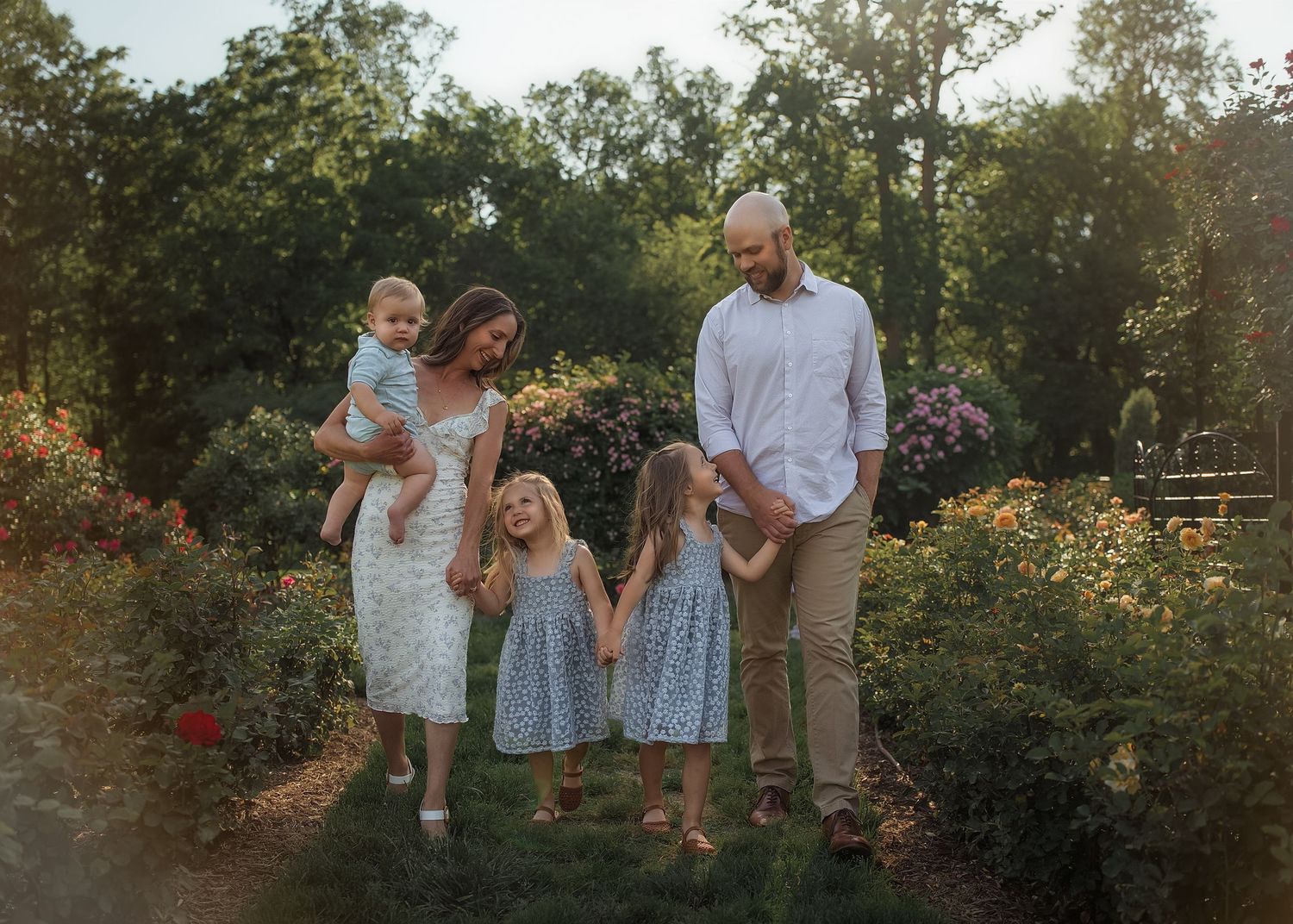 Family of five walking in a lush garden at golden hour, parents holding children in matching blue dresses.