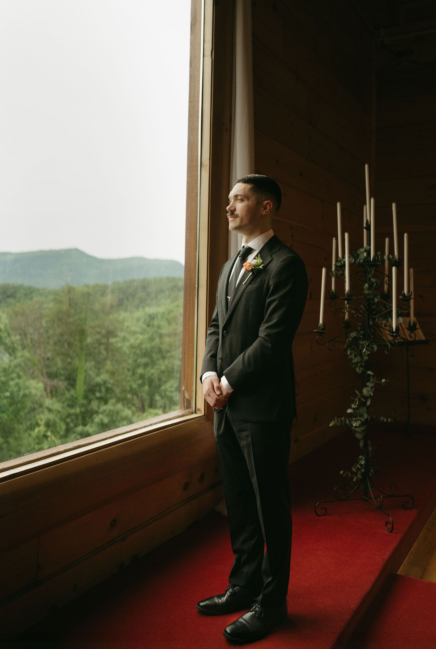 A formal suited figure gazes out a large window with candlesticks and mountain views.