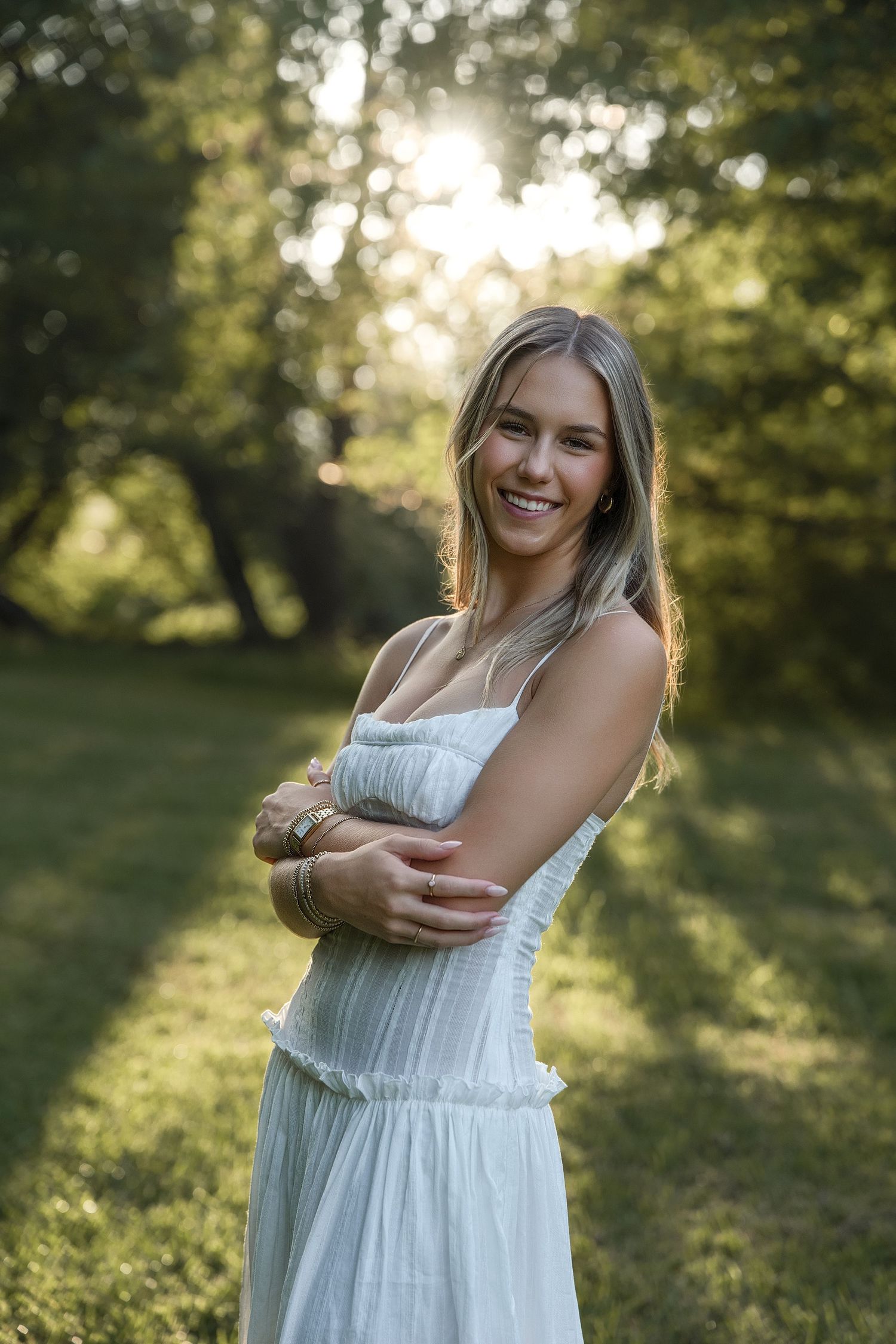 Smiling young woman in white sundress standing in sunlit green park, golden hour backlit portrait photography.