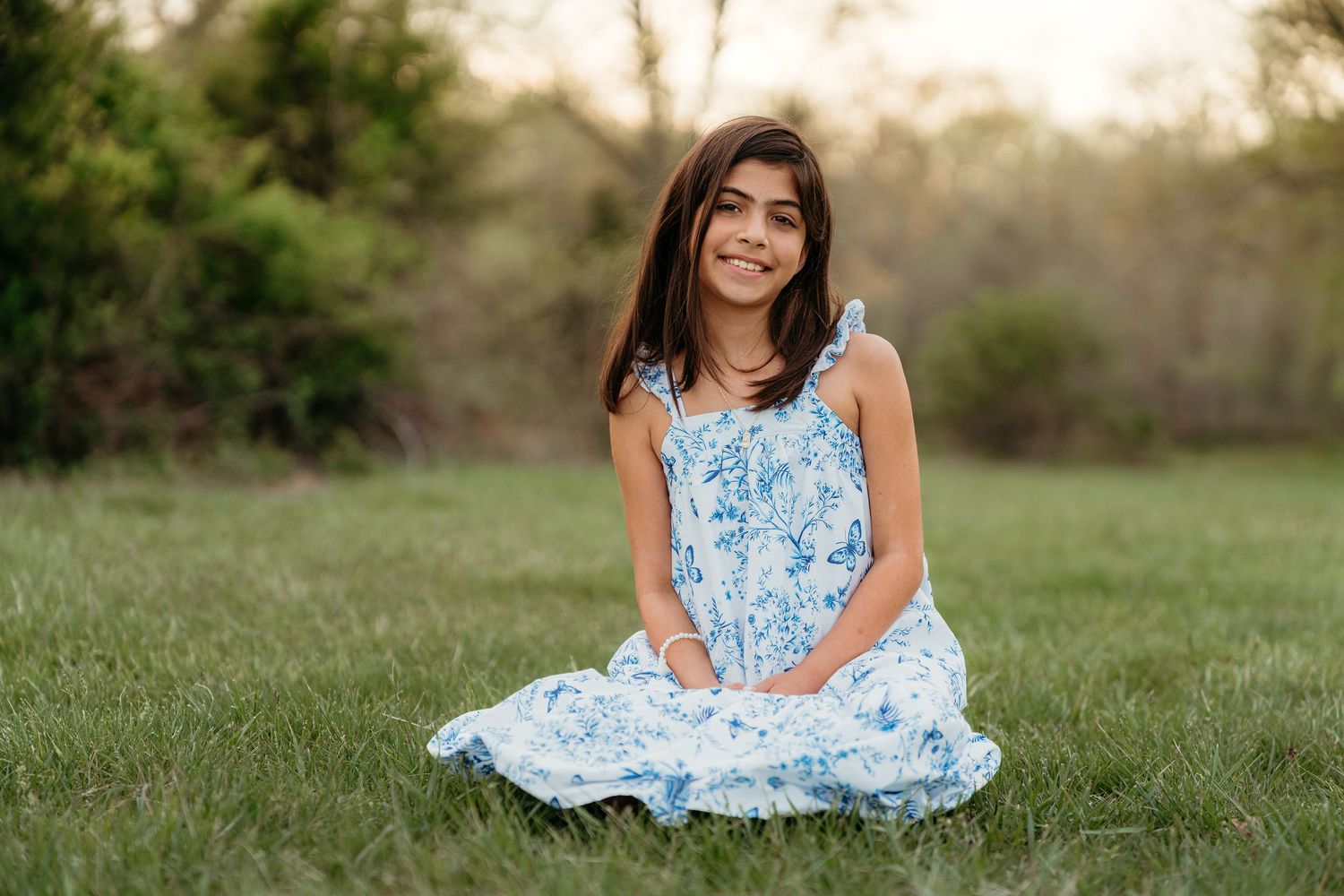 Gracefuly daughter in a floral dress sits gracefully on lush green grass near Haymarket Regional Lake during a sunny outdoor photo session.