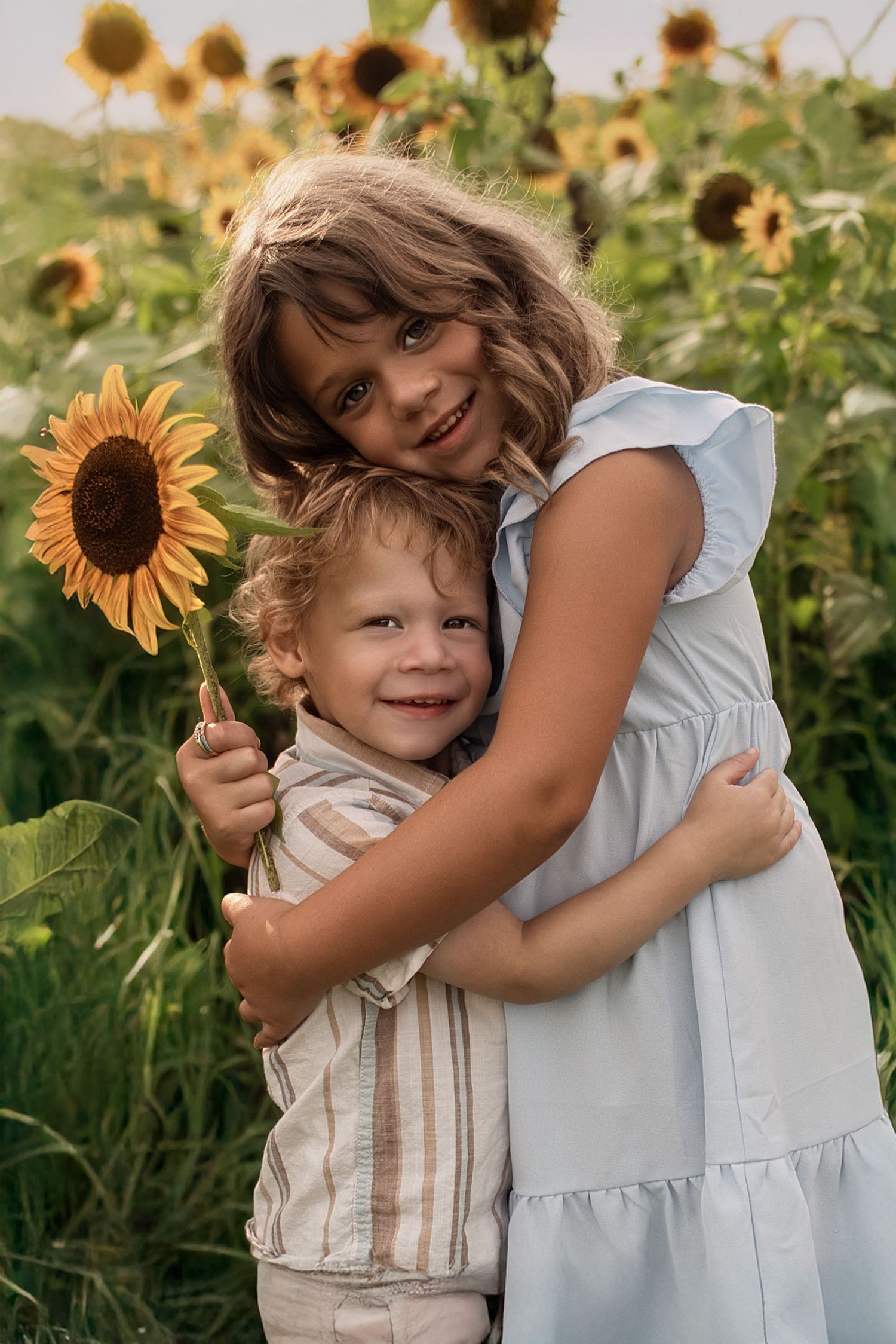 Children in white outfits share a tender moment among blooming sunflowers.