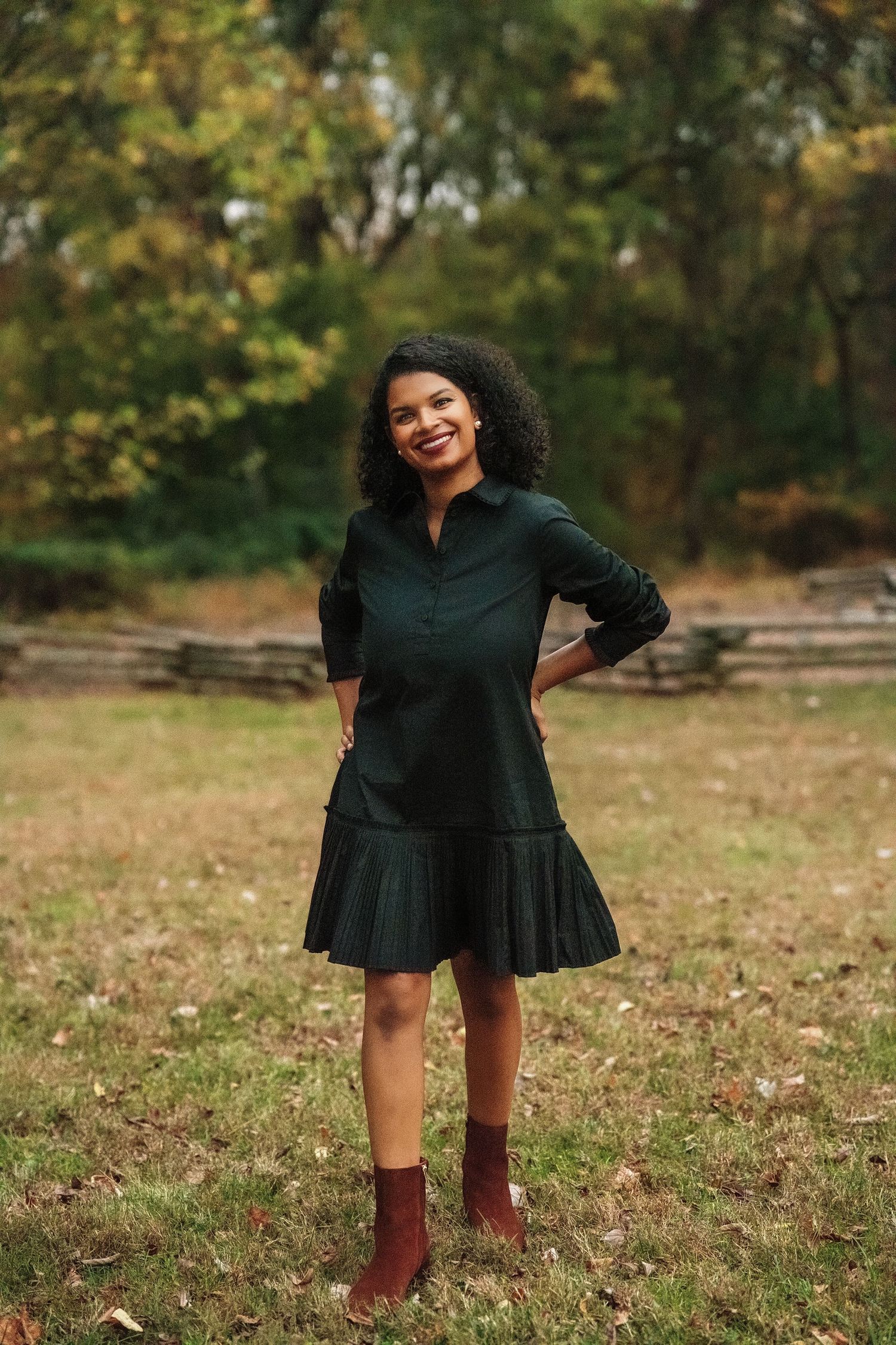 A person in a black dress and brown boots poses playfully on autumn grass in an outdoor setting.