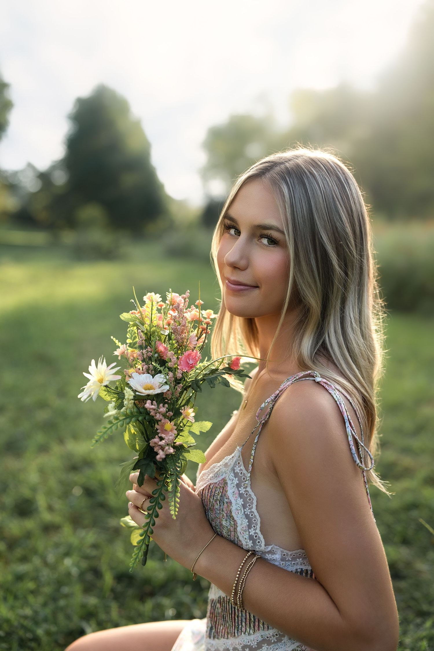 Young blonde woman holding a wildflower bouquet in a sunlit green field, wearing a lace dress and gold bracelets.