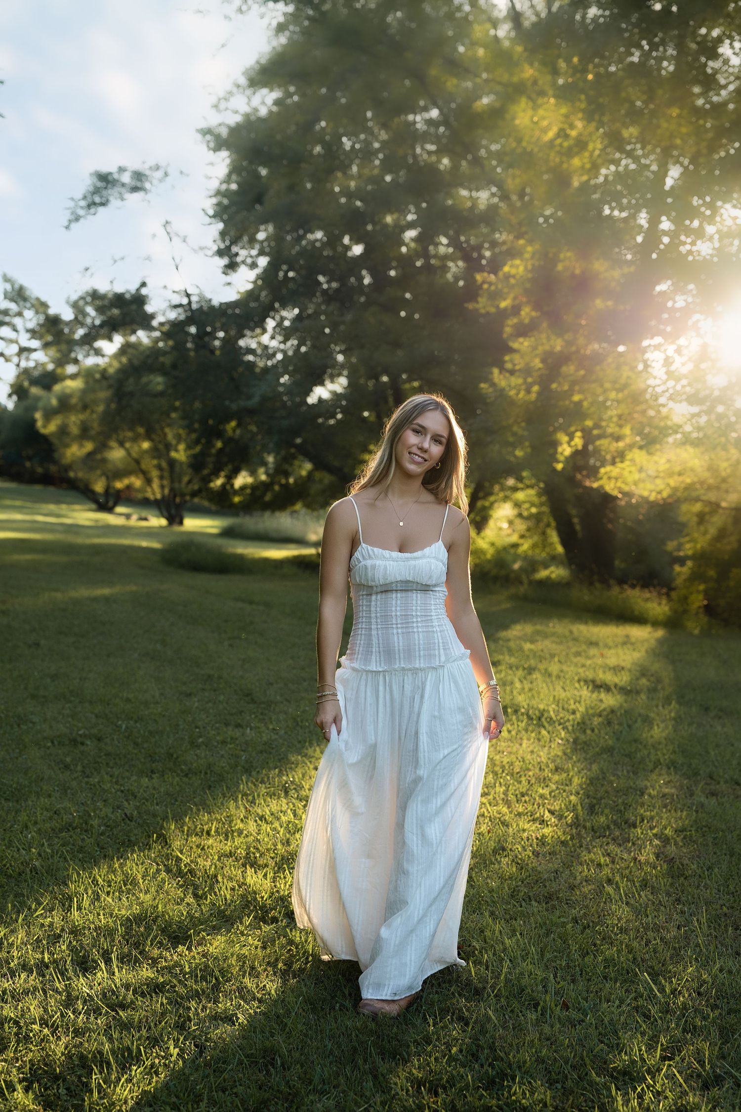 Young woman in white maxi dress walking through sunlit green park with trees glowing in golden hour light.