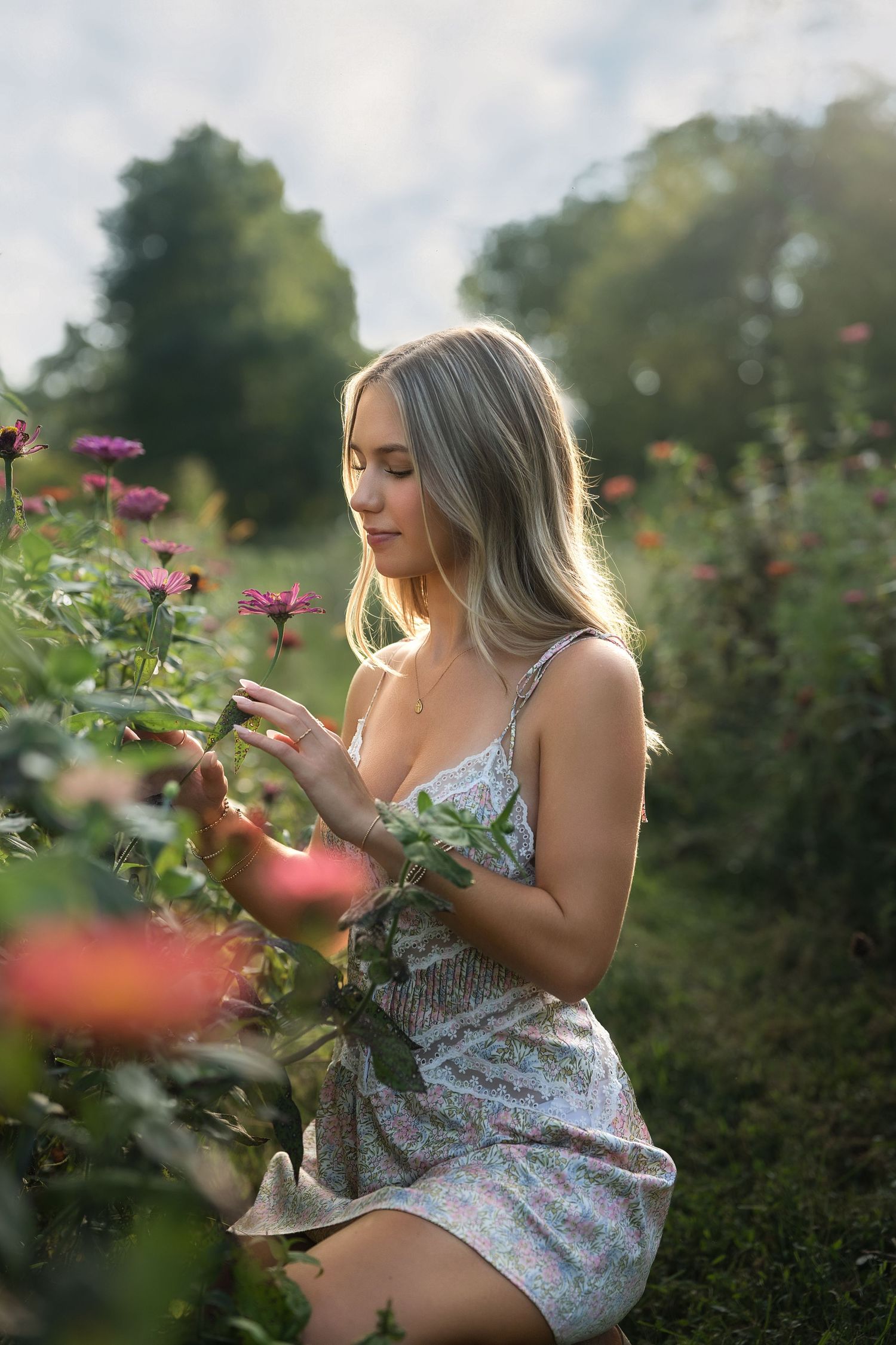 Young blonde woman in floral dress kneeling among pink zinnia flowers in a sunlit garden.
