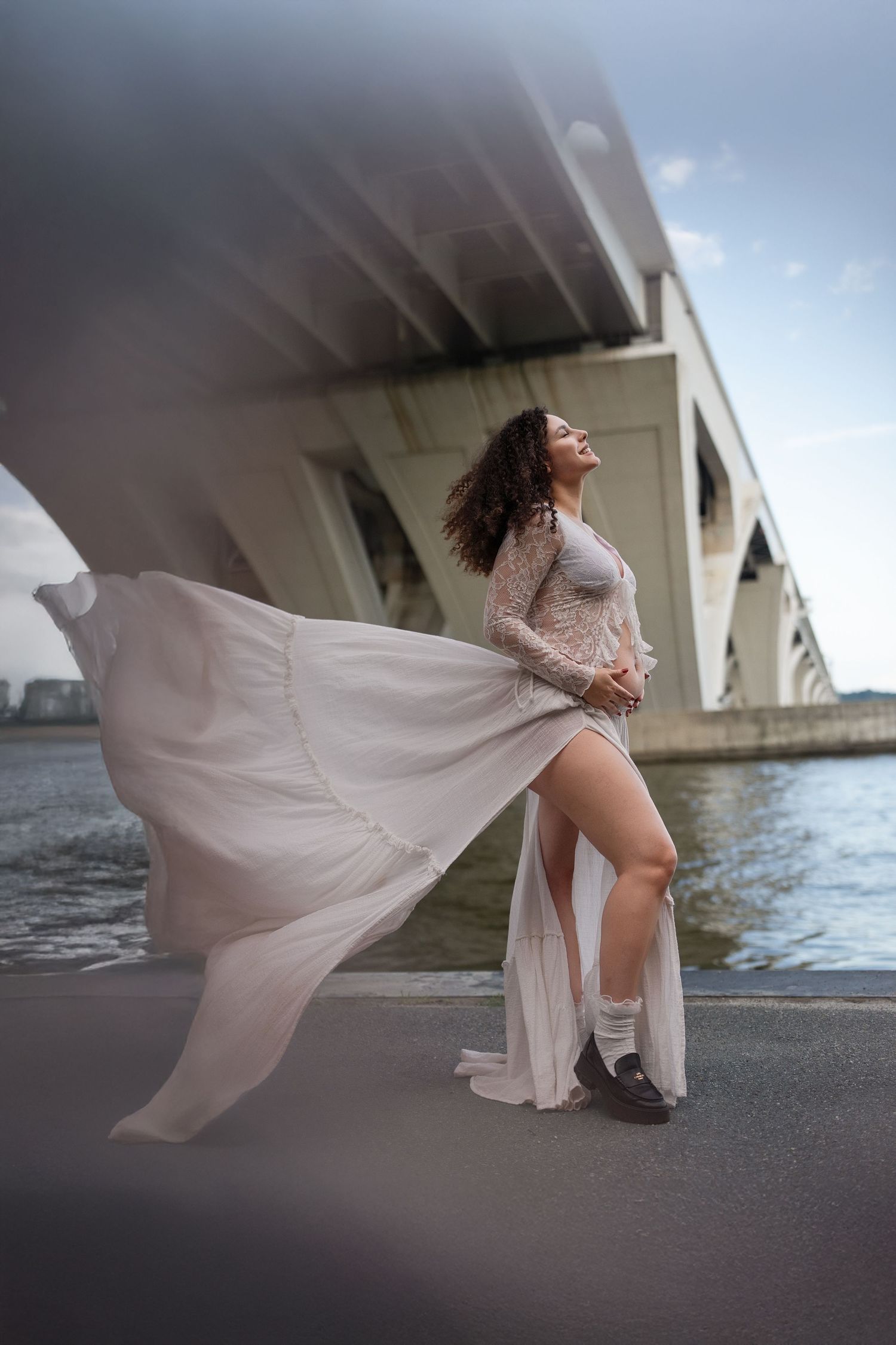 Dynamic maternity photos with billowing white dress under bridge architecture.