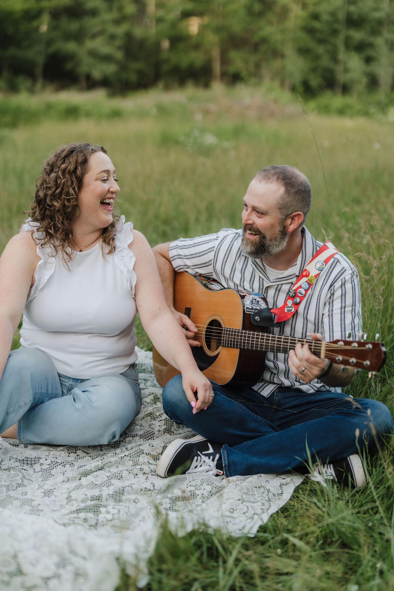A happy couple sits on a blanket in a green field, laughing together as the man plays an acoustic guitar.