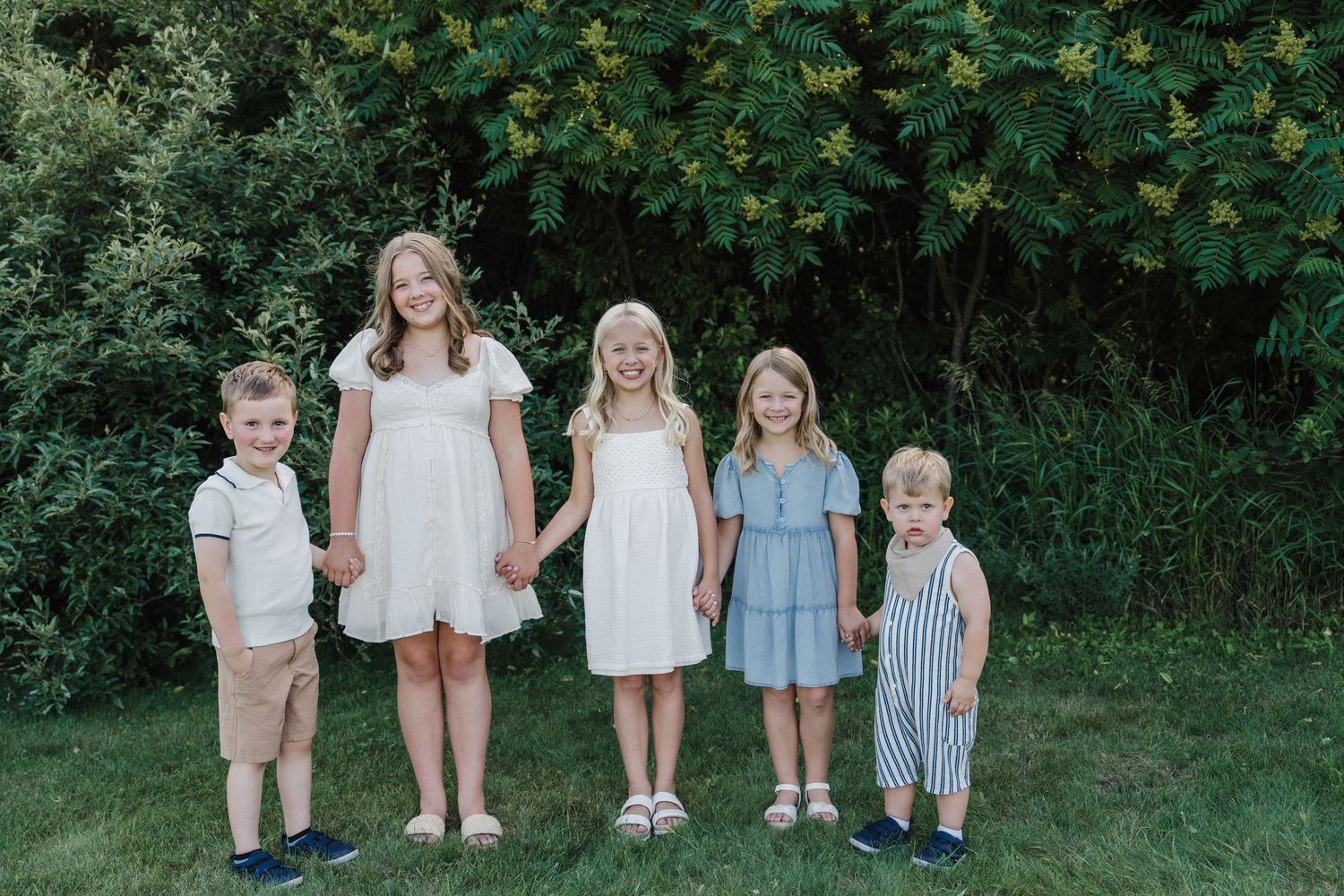 Five children in coordinated outfits pose holding hands on a green lawn in front of yellow flowering bushes.