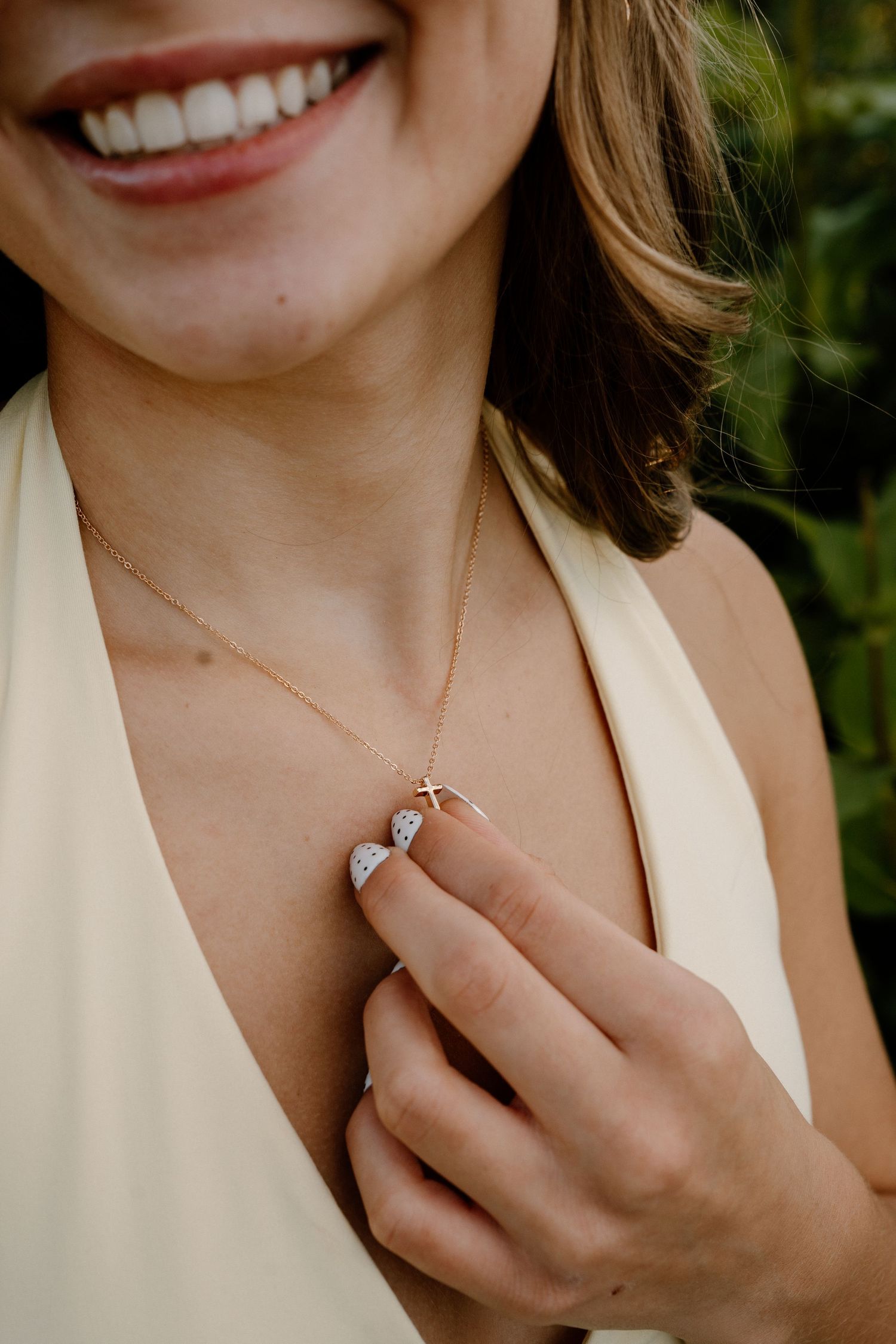 Woman smiling in white dress holding a delicate rose gold cross necklace with diamond heart pendant.