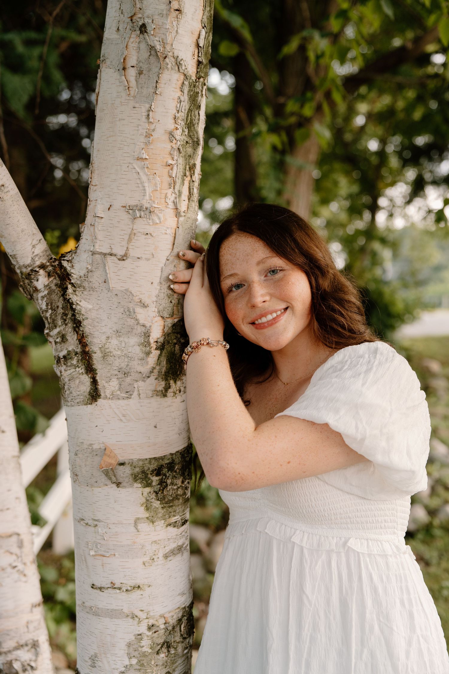 Young woman in white dress smiling while leaning against a birch tree outdoors.