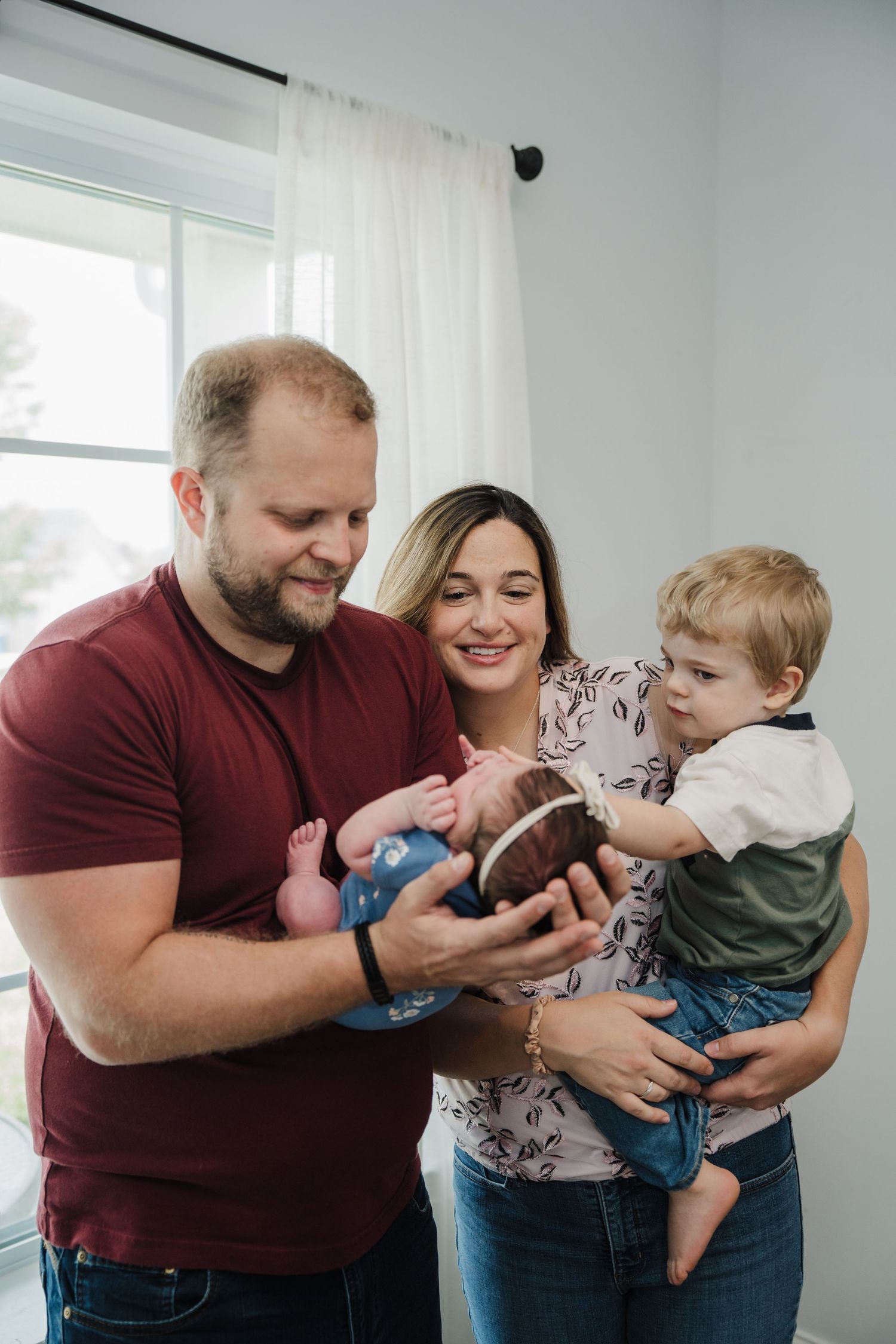 A happy family of four admires their newborn baby, with dad holding the infant and mom carrying their toddler son indoors.