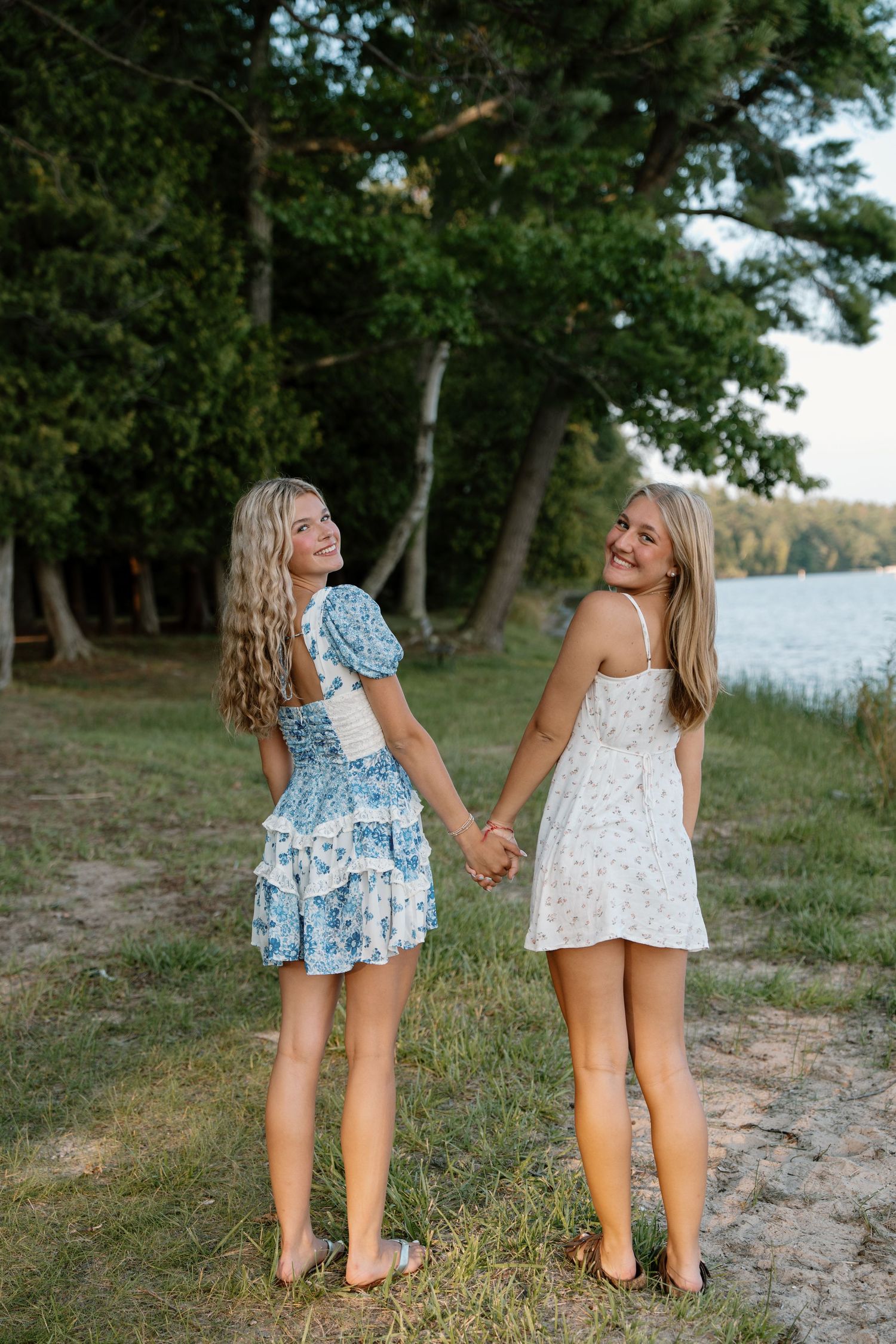 Two young women in floral summer dresses hold hands smiling near a lakeside tree-lined shore.