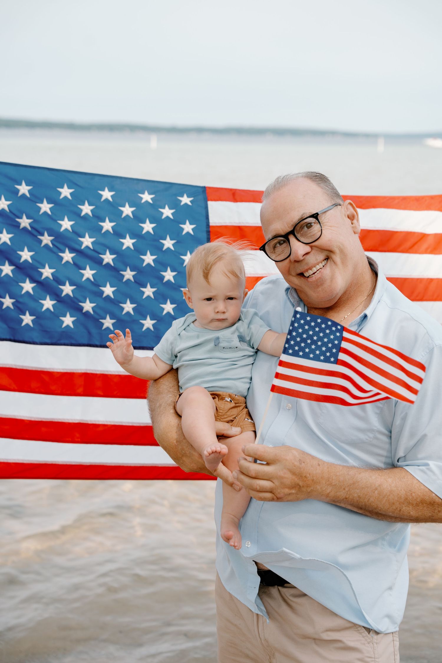 Smiling grandfather holding baby with American flags at the beach on Independence Day.