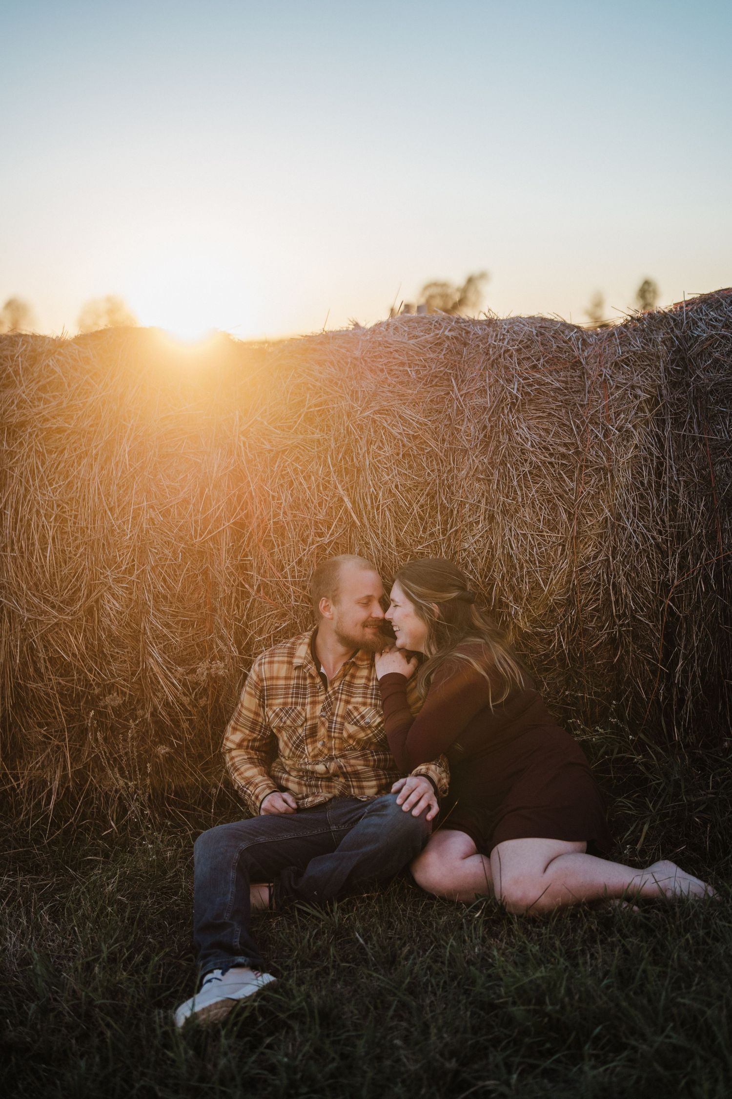 A couple lies together in a grassy field at sunset with golden light streaming through the background.
