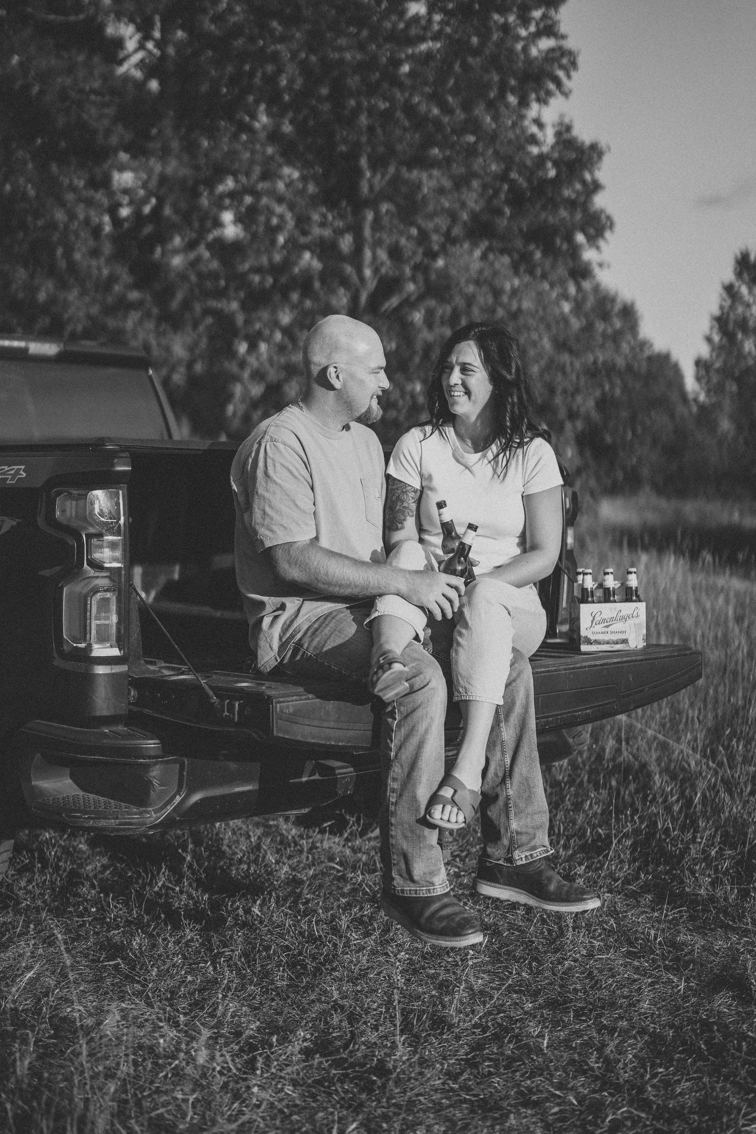 A couple sits together on the tailgate of a pickup truck sharing drinks in a casual outdoor black and white photo.