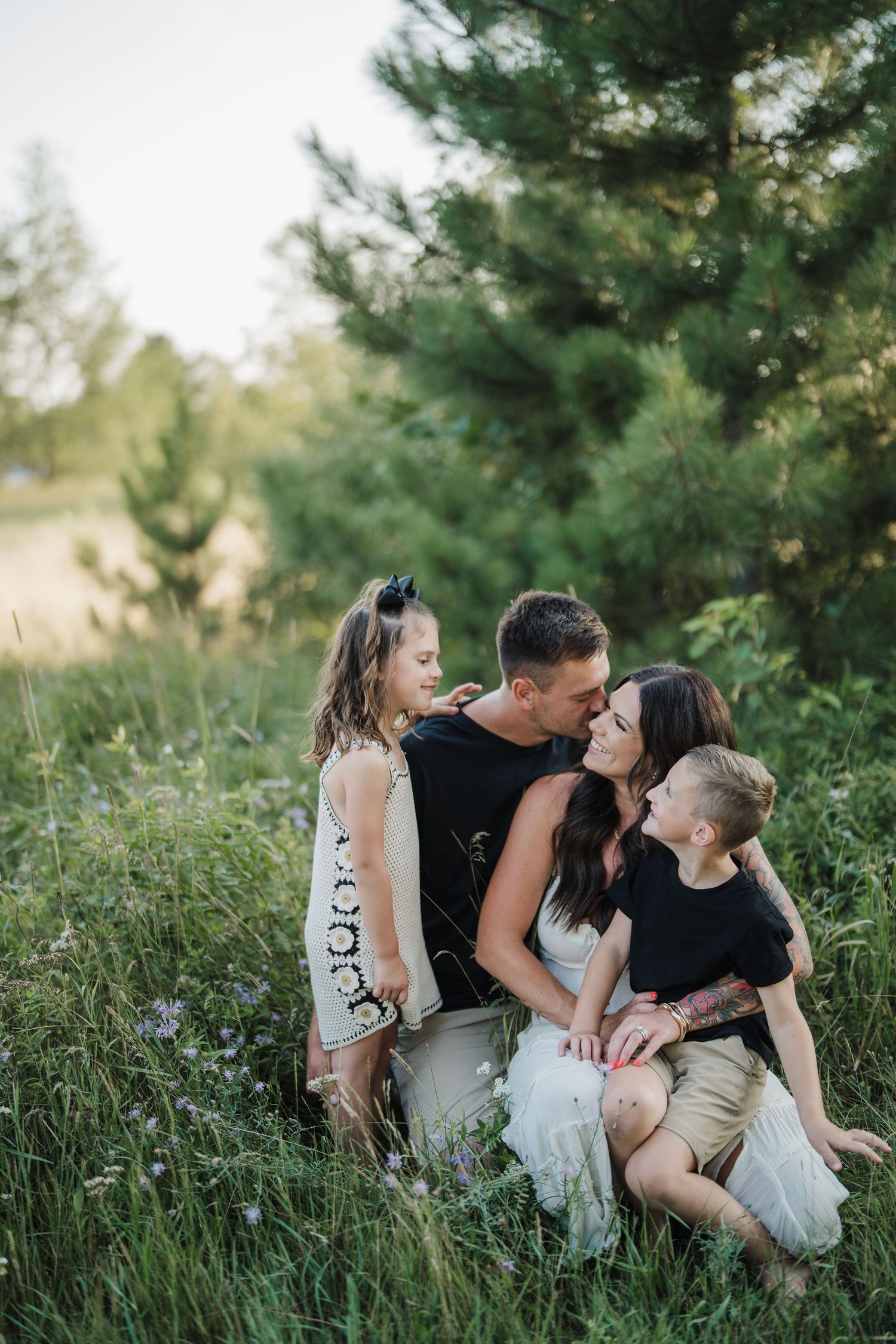 A romantic outdoor photoshoot in tall grass with soft golden light filtering through pine trees in the background.