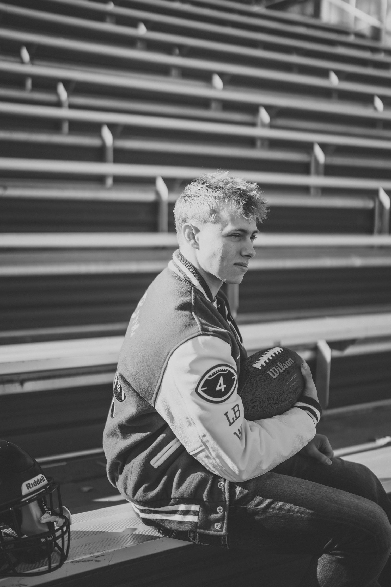 Black and white portrait of student sitting in stadium bleachers wearing varsity jacket.
