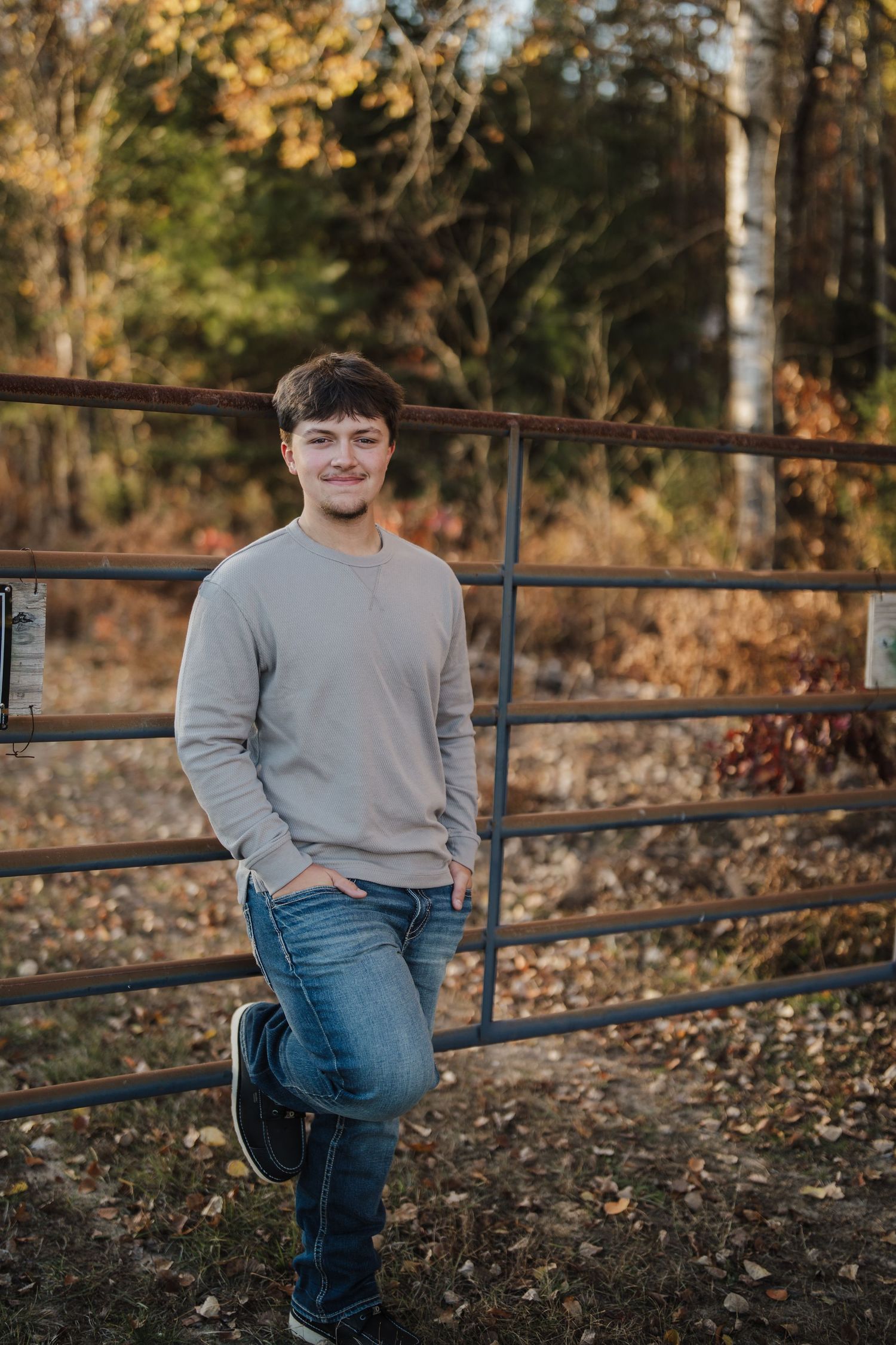 A person in a grey sweater and jeans leans casually against a rustic metal fence during golden hour.